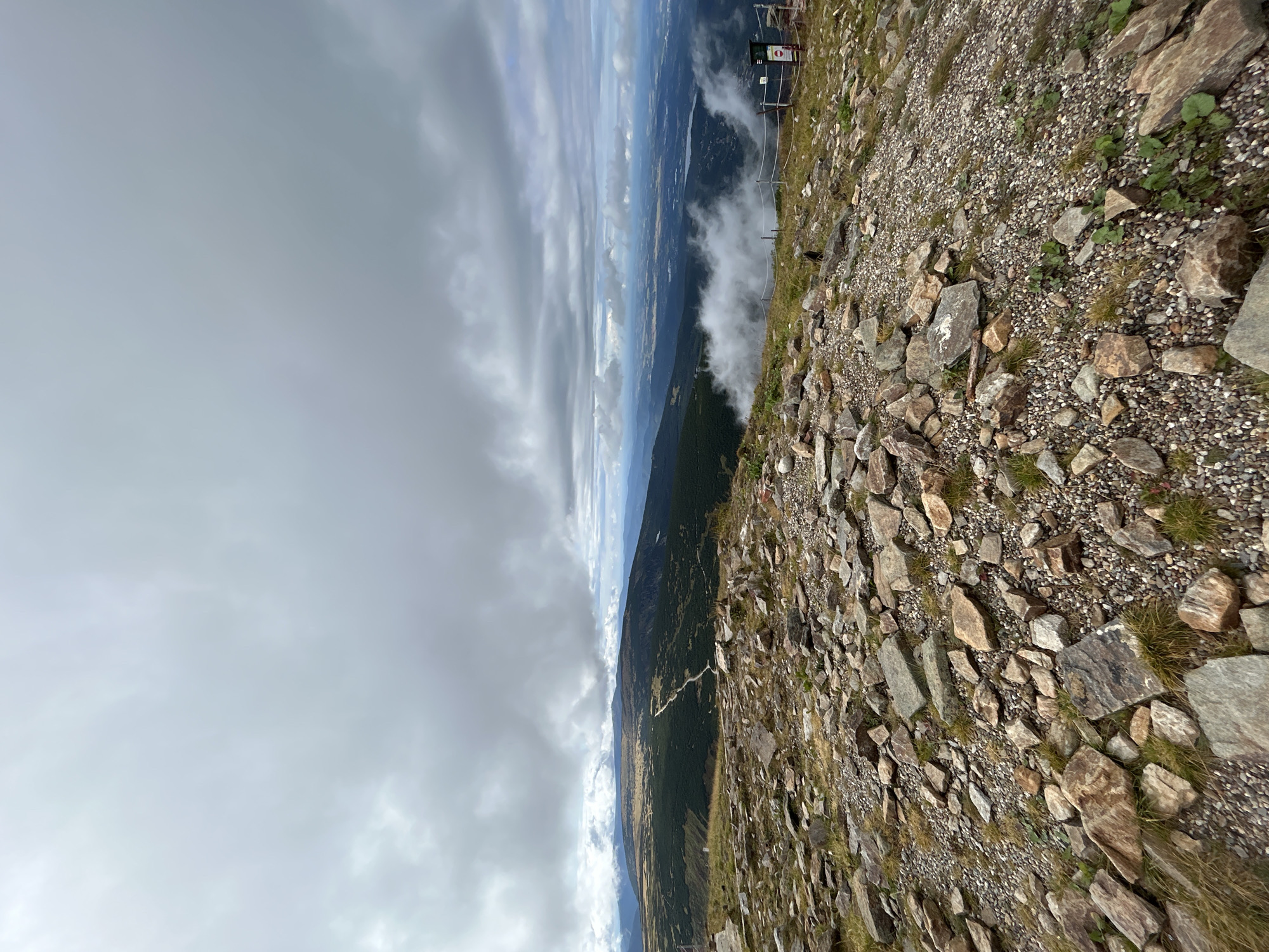 The image depicts a panoramic view from a rocky, elevated terrain overlooking a vast landscape. The foreground is dominated by scattered rocks of various sizes, some with visible striations, interspersed with patches of dry grass and small green plants. In the midground, a rolling green hillsides descends, marked by a winding path. Further in the distance, a valley unfolds with a body of water and scattered buildings. The sky is overcast with heavy, grey clouds, suggesting a cool, possibly damp, atmospheric condition. A wooden fence with a rope barrier runs along the edge of the terrain, with a small signpost visible on the right. The scene is likely a mountain area, given the elevation and rugged landscape. The presence of a path, fence, and signpost suggests it might be a tourist or hiking area.