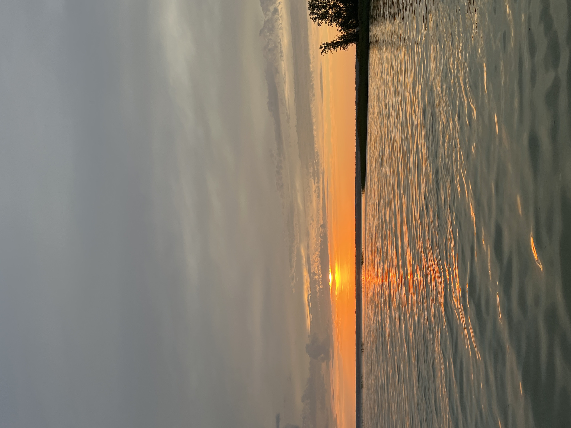 The image shows a serene lake at sunset, with the sun peeking through clouds on the horizon, casting an orange glow across the water. The sky is a mix of gray and orange hues, with streaks of clouds. The water has gentle ripples, reflecting the colors of the sunset. A distant treeline marks the edge of the lake, with a few trees visible on the right side of the frame. The scene appears to be in a natural, tranquil setting, potentially in Beregovoe, Russia, a coastal village known for its scenic beauty. There are no people or specific objects depicted that would suggest a particular activity or interaction. The overall atmosphere is one of peacefulness and natural beauty.