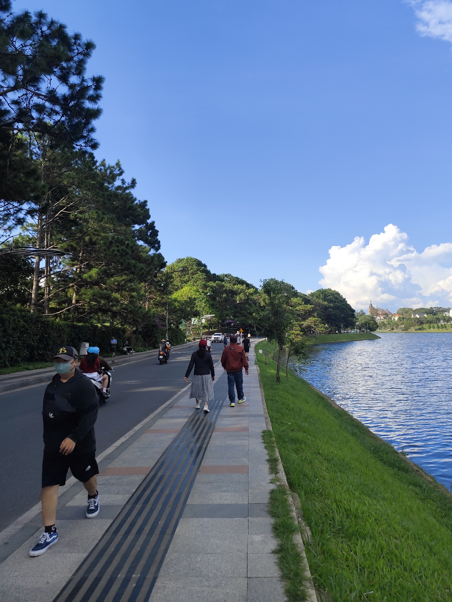 The image depicts Xuan Huong Lake in Da Lat, Vietnam, on a bright, sunny day with a clear blue sky and scattered white clouds. A paved pathway runs along the edge of a road, bordered by a grassy embankment that slopes down to the lake. Several people are walking on the pathway. In the foreground, a person wearing a black hoodie, black shorts, a face mask, and blue sneakers walks towards the camera. Further down the path, another person in a black jacket and grey skirt walks away from the camera, followed by a person in a red jacket. Motorcyclists are visible on the road. Pine trees and other lush green vegetation line the opposite side of the road and extend up a gentle incline where buildings are visible in the distance. The lake's surface is a vibrant blue, reflecting the sky and creating a serene atmosphere.