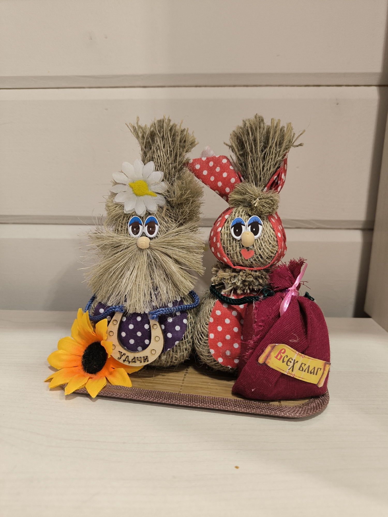 The image shows two handmade stuffed bunny figures made of burlap and straw, decorated with fabric accents and accessories. The bunny on the left has a white daisy on its head and a purple polka-dot apron with a horseshoe charm that reads "Удачи" (Good luck). It is accompanied by a yellow artificial sunflower. The bunny on the right wears a red polka-dot headscarf and dress, and is holding a small burgundy sack tied with pink ribbon, with a yellow tag that reads "Всех благ" (All the best). Both figures have button eyes and felt mouths. They are positioned on a woven mat, which is on a light-colored wooden surface. The background consists of a light-colored, horizontally paneled wall. The scene is indoors, likely on a shelf or table, and appears to be a decorative display. There are no people or animals present. The image does not contain any specific information or visual cues that would place it within the city of Oktyabrsky, Russia.