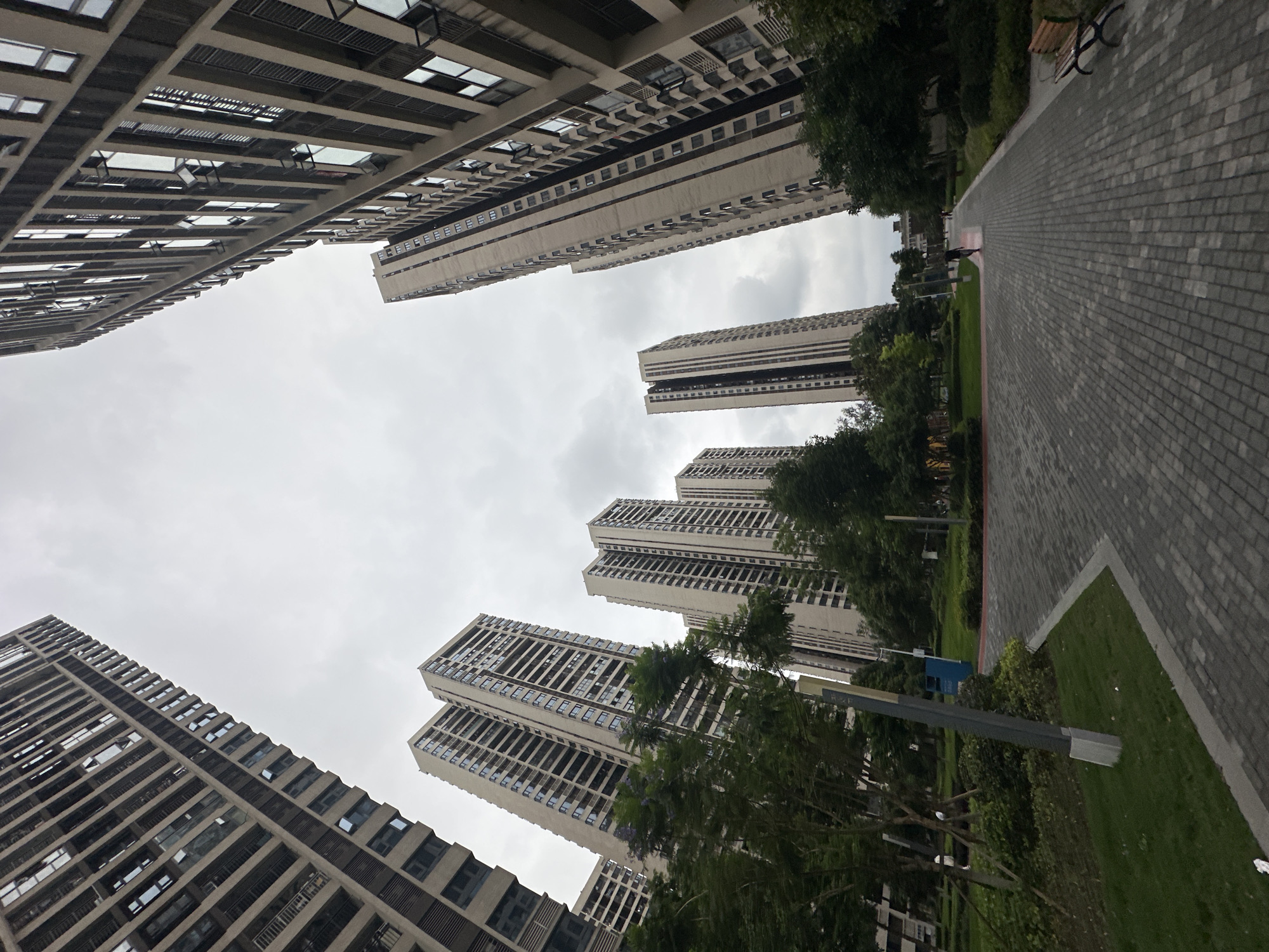 The image depicts a view of multiple high-rise apartment buildings in Chongqing High-tech Zone, China. The buildings are a light grey color and feature multiple windows. The sky is overcast with grey clouds. In the foreground, a pedestrian walkway made of light grey bricks is visible, with green grass and trees bordering it. A light pole stands alongside the walkway.