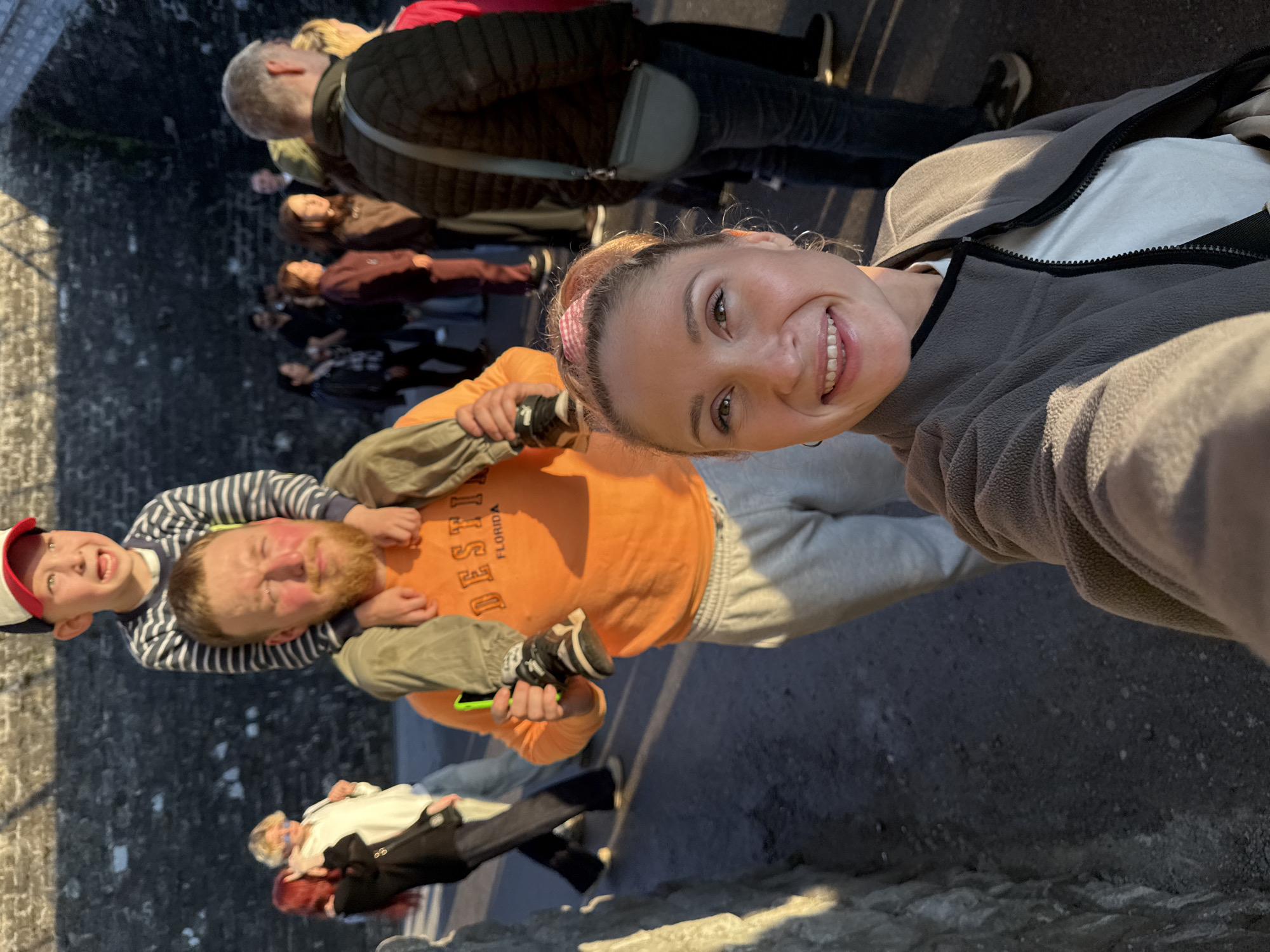 This image captures a group of people outdoors, possibly in a city. In the foreground, a woman is taking a selfie, smiling directly at the camera. She is wearing a gray fleece jacket over a white t-shirt. Behind her, a man with a beard is holding a young boy on his shoulders. The boy is wearing a striped shirt and a baseball cap, and appears to be shouting or making a face. The man is wearing an orange sweatshirt with the word "DESTINY FLORIDA" visible on it, and gray sweatpants. He is holding a bright green object in his left hand.
In the background, a crowd of people are visible, some of whom are walking. The environment appears to be a paved area with a textured stone wall on the left side of the image. The lighting suggests it is either early morning or late afternoon, with a warm glow.
While the image does not explicitly show any identifiable landmarks or street signs, the presence of a textured stone wall and the general urban setting could be consistent with various locations. Without further information or context, it is not possible to definitively place this scene within the city of Perm, Russia, or any other specific city.