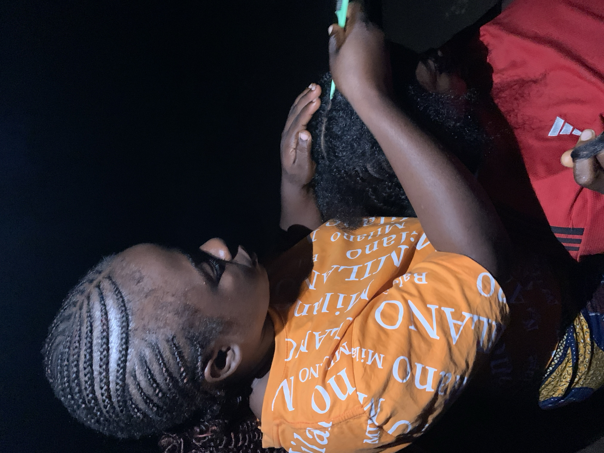 In Akwanga, Nigeria, a close-up shot focuses on two individuals engaged in hairstyling. The primary subject, positioned on the left, is a woman with intricately braided hair, her head tilted as she concentrates on her task. She is wearing an orange t-shirt emblazoned with white lettering that appears to be a repeating pattern. To her right, another individual, whose face is largely obscured by shadow and the action, is having their hair styled. The stylist's hands are visible, one holding a green comb and the other sectioning hair. The background is dark, suggesting an indoor setting or nighttime conditions, with minimal environmental detail visible. The lighting emphasizes the people and the hair, creating a dramatic contrast with the surrounding darkness.