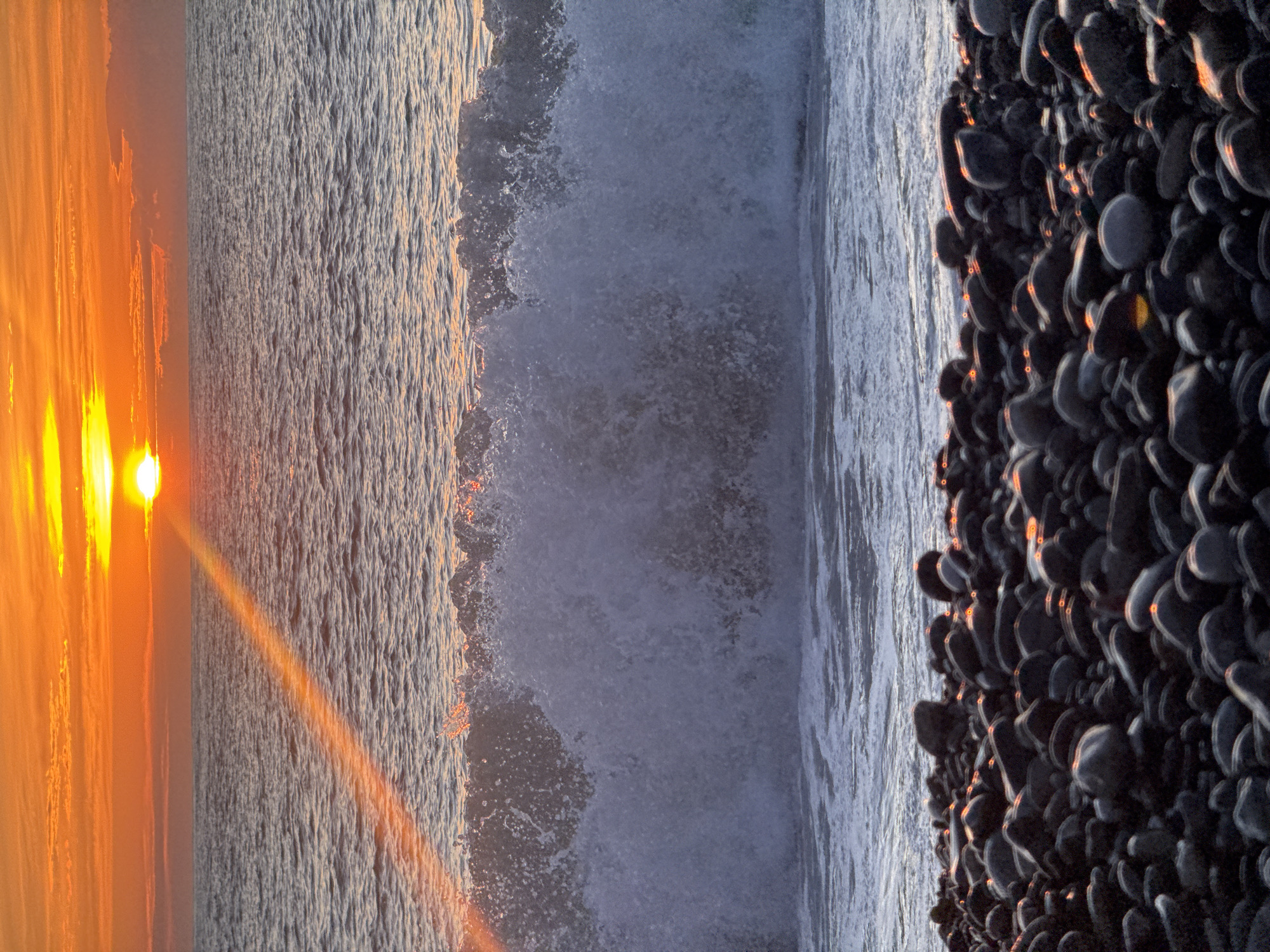 The image captures a vertical view of a beach scene, likely at sunset. The left side of the image displays a bright orange sunset with the sun visible. Adjacent to this is the ocean, where waves are crashing. On the right, a pile of dark, wet pebbles is seen. The overall atmosphere suggests a twilight setting on a beach. It's difficult to confirm the location is Batumi, Georgia without additional context clues from the scene.