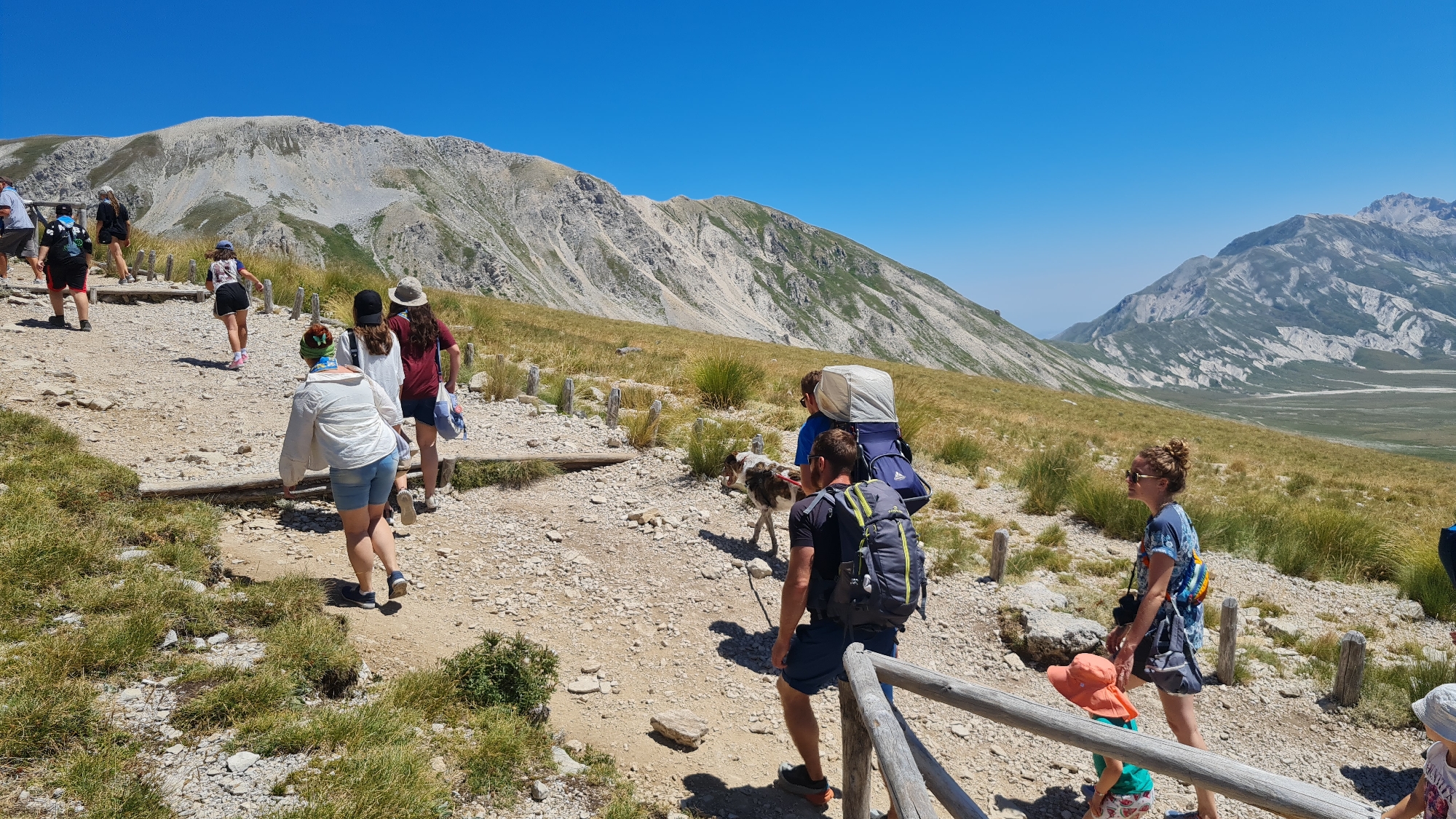 This image depicts a group of people and a dog hiking on a mountain path under a clear blue sky. The path is made of gravel and stones, with some grassy areas and wooden posts lining the sides. In the background, there are rolling hills and large mountains with rocky slopes.
There are several individuals visible, most of whom appear to be walking uphill. One person in the foreground is wearing a white jacket and denim shorts, with a colorful bandana on their head. To their right, a woman in a blue patterned top and shorts is walking with sunglasses on. A child in an orange hat and green shirt is walking ahead of her. To the left of the woman in white, there are two people walking together, one in a dark shirt and shorts with a backpack, and another in a blue shirt carrying a large pack. A dog with brown and white markings is walking alongside them. Further up the path, several other people are visible, some with backpacks.
The environment suggests a mountainous, rural location, possibly a national park or hiking trail. The presence of wooden posts and constructed path elements indicates a maintained hiking route. The bright sunlight and clear sky suggest a warm, summer day. While the specific location in L'Aquila, Italy, is not explicitly shown, the landscape is consistent with the mountainous terrain found in the Abruzzo region surrounding L'Aquila, known for its national parks and outdoor recreational activities.