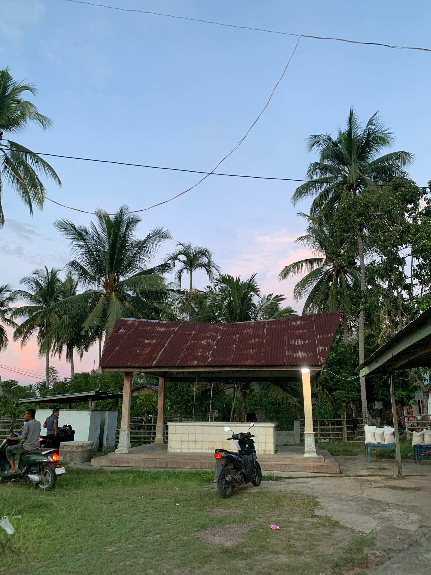 The image depicts an outdoor scene at dusk, likely in the city of Palohawe, Indonesia, characterized by lush palm trees and a soft twilight sky with hues of pink and blue. In the foreground, a grassy area leads to a paved pathway. To the left, two men are on a dark green scooter, with one appearing to drive and the other as a passenger. A black motorcycle is parked in the center of the frame.
Dominating the midground is a structure with a weathered, corrugated metal roof supported by columns. This structure appears to be a covered pavilion or a small shrine, with a tiled area within. A bright light emanates from one of the columns. Beyond this structure, a wooden fence is visible. To the right, a low-lying building with a similar roofline and a table holding several sacks of what appear to be rice or grain. The background is filled with dense foliage and more palm trees, indicative of a tropical environment. The overall impression is one of a rural or semi-rural setting during the transition from day to night.