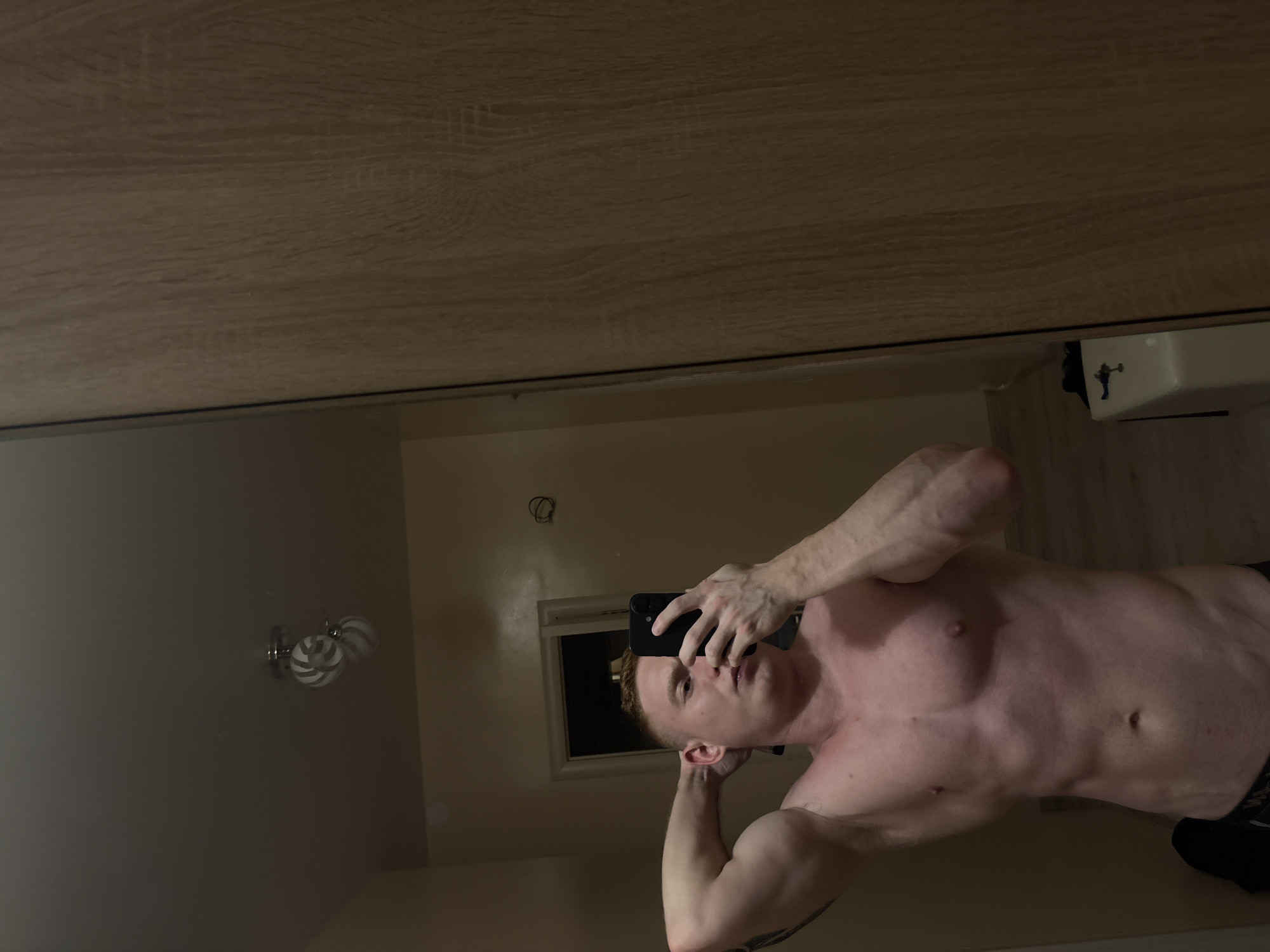 A shirtless, light-skinned male stands in a bathroom with light-colored walls and a light wood-toned ceiling, taking a selfie in a mirror. He is holding a black smartphone in his right hand, obscuring part of his face. His left arm is raised, hand near his head. A three-bulb vanity light fixture is visible on the wall to his left. A white sink and wall-mounted fixture are visible in the upper right of the mirror. The room is dimly lit.
There is no verifiable information to indicate that this photo was taken in Chaikovsky, Russia.