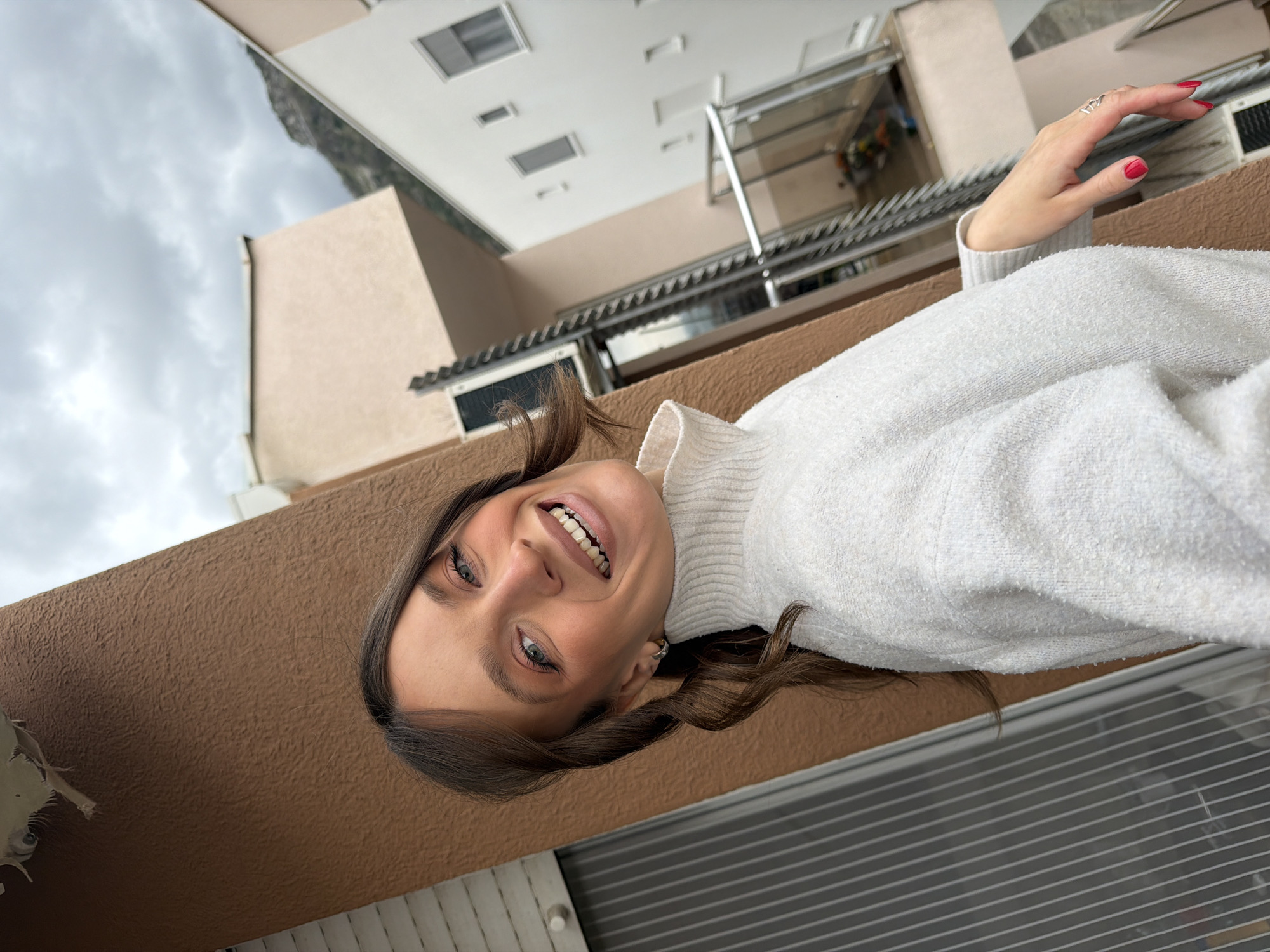 A young adult female is depicted in a close-up, angled portrait, positioned outdoors on what appears to be a balcony or terrace in Budva, Montenegro. She has long, wavy brown hair and blue eyes, and is smiling broadly while looking directly at the camera. She wears a cream-colored, textured turtleneck sweater. Her left hand is visible, featuring red nail polish and a silver ring on her left ring finger. The immediate foreground includes a brown, textured stucco wall or pillar to her left. The background consists of residential buildings; a white building with multiple dark-framed windows is prominent to her right, and another light-colored building with an external air conditioning unit is partially visible behind her. Below the subject's right arm, a lower balcony with a glass and metal railing contains a visible pot with flowers. A mountainous landscape under a heavily overcast sky forms the distant background, establishing the setting in Budva, Montenegro.