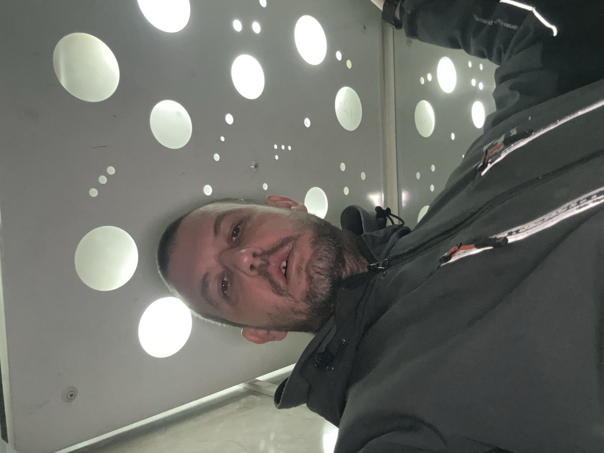 A man with a short haircut and beard is looking down at the camera. He is wearing a dark jacket with white accents. Behind him is a wall with numerous round, illuminated holes. The location appears to be an enclosed space, possibly an elevator or a small room with specialized lighting. The city is Sofia, Bulgaria, but there are no identifying markers in the photo that definitively prove the location.