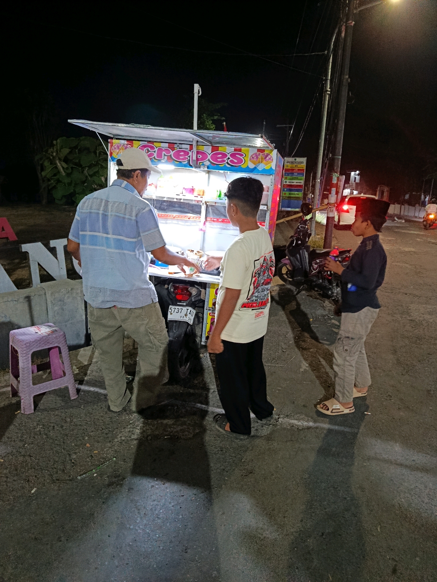 At night in Sumber Suko, Indonesia, a man wearing a blue and white striped short-sleeved shirt, khaki cargo pants, and a light-colored baseball cap stands behind a crepe stand. He appears to be preparing food. Two young boys are present. One boy, wearing a white t-shirt with a graphic print and black pants, stands to the man's right, looking at the stand. The other boy, wearing a dark long-sleeved shirt and light gray cargo pants, stands to the far right, holding a phone. A purple stool is to the left of the man. A motorcycle is parked behind the crepe stand. The ground is asphalt, with some debris, including cigarette butts, scattered on it. A large concrete structure with white lettering, partially obscured, is visible in the background to the left, along with some vegetation. A street light illuminates the scene from the right, casting long shadows. A red brake light is visible on a vehicle in the distance.