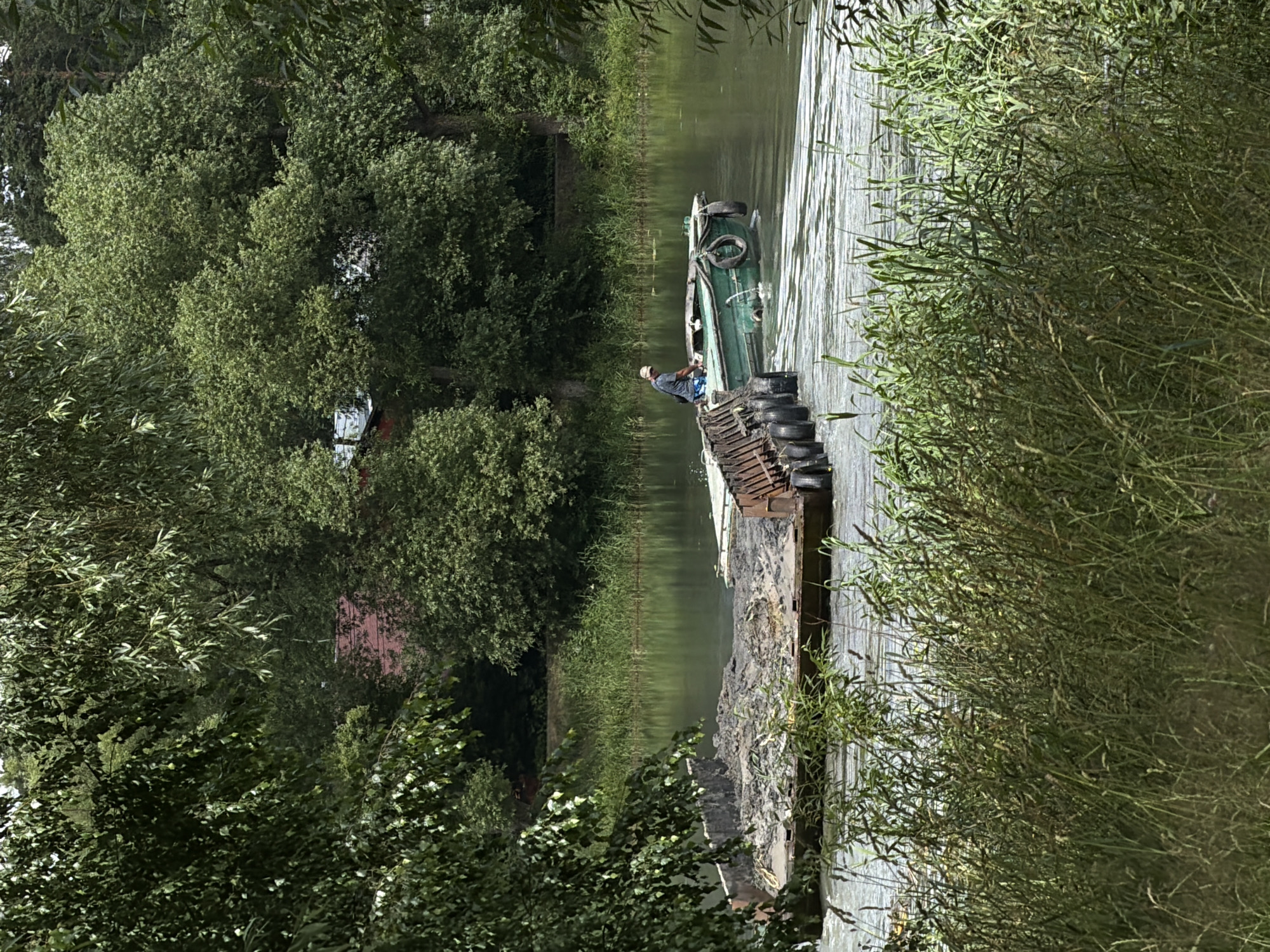 In Bykovo, Russia, on a body of water that appears to be a river or canal, a person is operating a small green boat. The boat is moving alongside a large, flat barge loaded with dark material, possibly soil or construction debris. The barge is equipped with numerous black tires along its side, serving as fenders. The environment is lush and green, with dense trees framing the scene from the left and in the background. A red-roofed building is visible through the trees in the distance. The water is calm, with ripples indicating the movement of the boats. Tall grasses and reeds grow along the water's edge in the foreground. The sky is overcast.