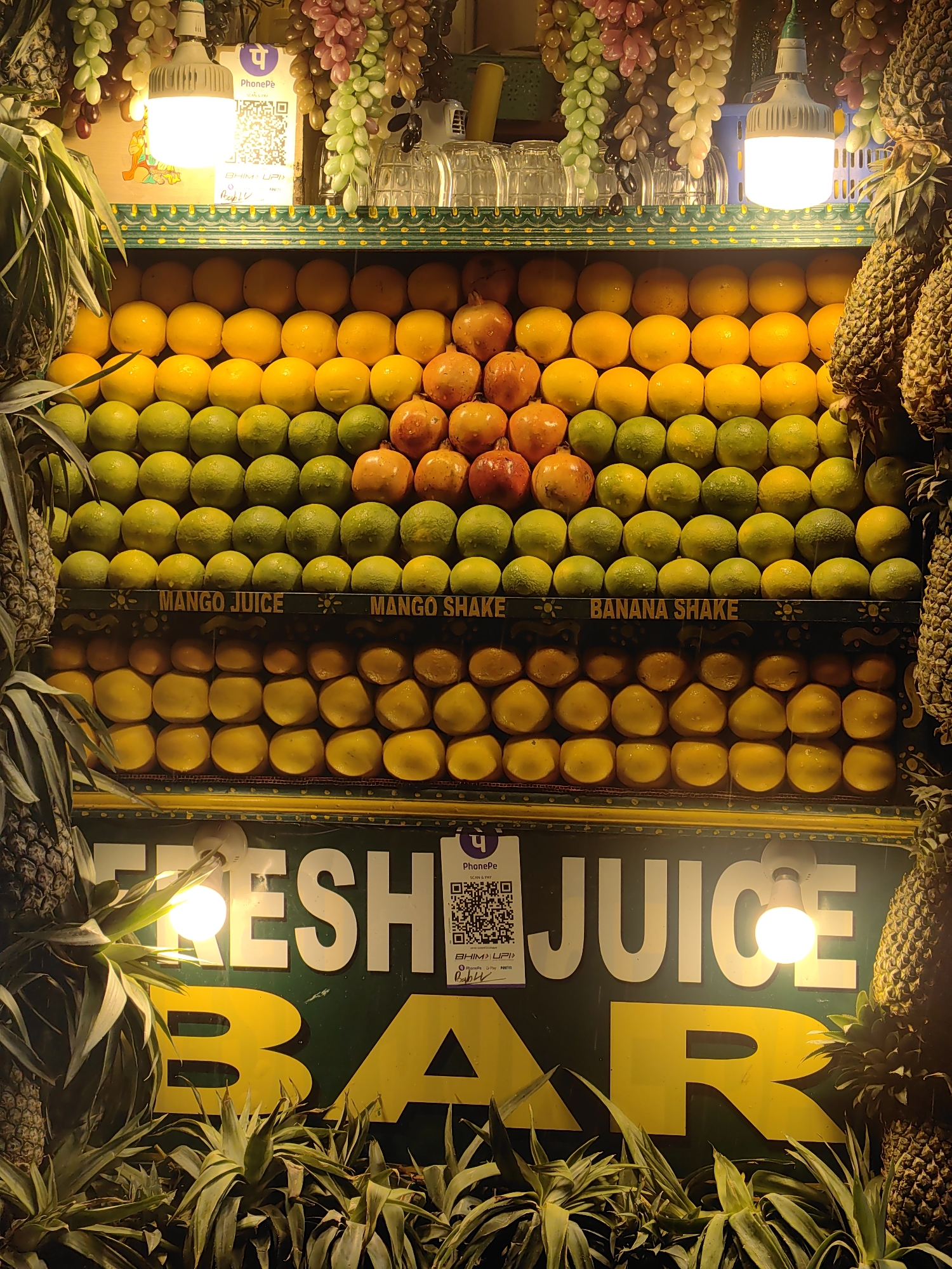 A brightly lit juice bar in Manali, India, displays a vibrant assortment of fresh fruits meticulously arranged on shelves. Oranges, lemons, and pomegranates are stacked in neat rows, with pineapples flanking the display. The bar's signage, in bold white letters against a dark green background, proclaims "FRESH JUICE BAR." A QR code for PhonePe, a digital payment service, is visible, alongside decorative strings of artificial grapes and glasses displayed above the fruit. The scene is captured at eye level, creating an inviting and eye-catching presentation of the available offerings.