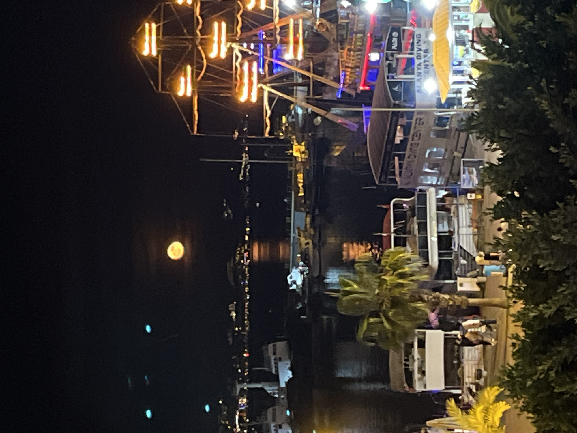 The image, originally oriented sideways, captures a vibrant night scene along a waterfront in Alanya, Turkey. A large, bright full moon illuminates the dark sky on the left side of the frame, casting a soft glow over the water below.
On the right, a lively pier or marina is bustling with activity. Several boats are docked, many elaborately decorated with artificial lights that reflect on the water's surface. A particularly striking feature is a large, cage-like structure adorned with numerous warm, golden-orange vertical lights, possibly part of a tour boat or a fixed installation. Other boats display colorful blue and red illuminations. One prominent vessel clearly advertises "ALANYA DIVING CENTER" and "PADI" on its side, suggesting a recreational and tourist-focused environment.
Green trees and palm trees, with some yellow-lit foliage, frame the right foreground, partially obscuring parts of the waterfront. Indistinct figures of people can be seen moving about on the boats and along the pier, indicating a social atmosphere. The overall impression is one of an energetic night out in a coastal town, with the moon overseeing the illuminated harbor activities.