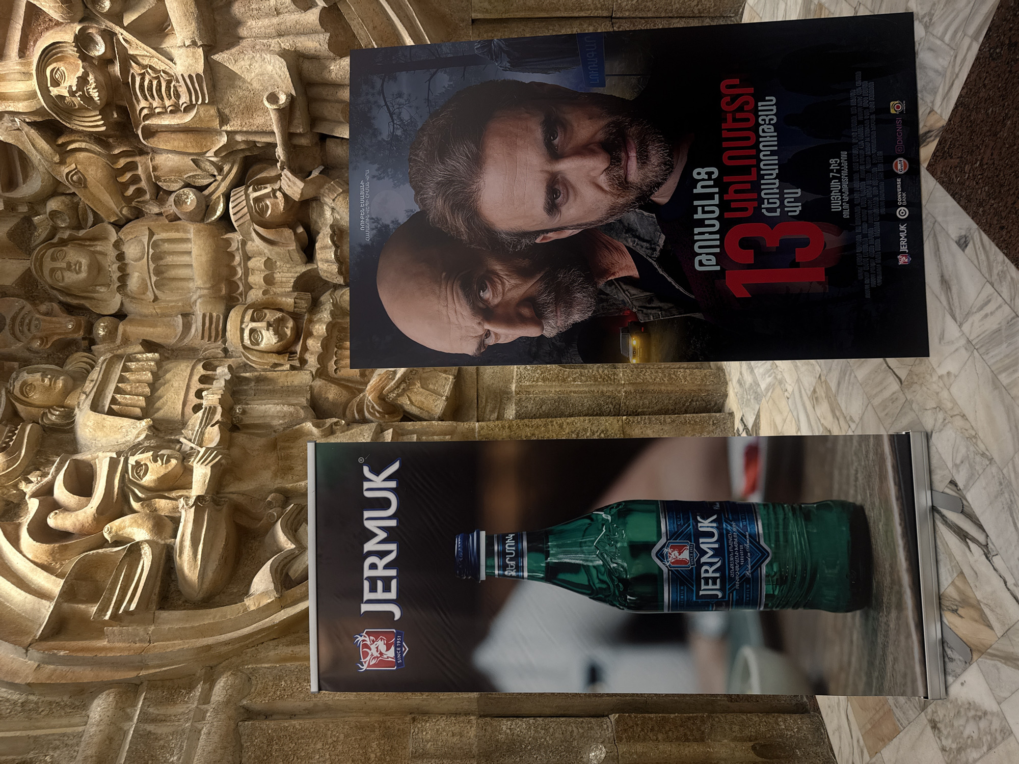 The image captures a display near a wall in Yerevan, Armenia. The wall is intricately carved with figures in relief. Two posters are prominently displayed. The poster on top is a movie ad for a film titled "13 Kilometers," featuring the faces of two men. The lower poster is an advertisement for Jermuk mineral water, displaying a green bottle of the product. Both the movie title and brand name are printed in Armenian script as well. The background is a marble pattern, likely the floor.
