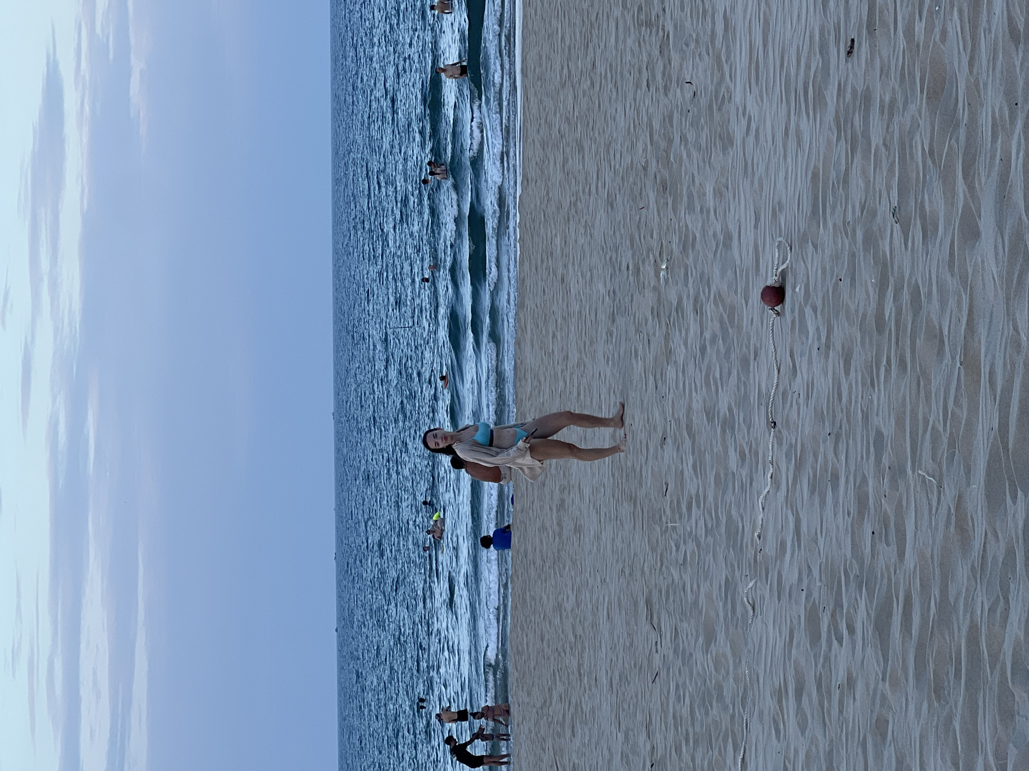 A young woman in a blue bikini and a tan cover-up stands on a sandy beach in Da Nang, Vietnam. She is facing the camera and looking directly at it. The ocean is behind her, with several people swimming and playing in the water. The sky is a pale blue with some clouds. There is a red buoy and rope on the sand in the foreground. The beach is populated by several other people, some of whom are interacting with each other. The overall scene is one of a casual day at the beach.