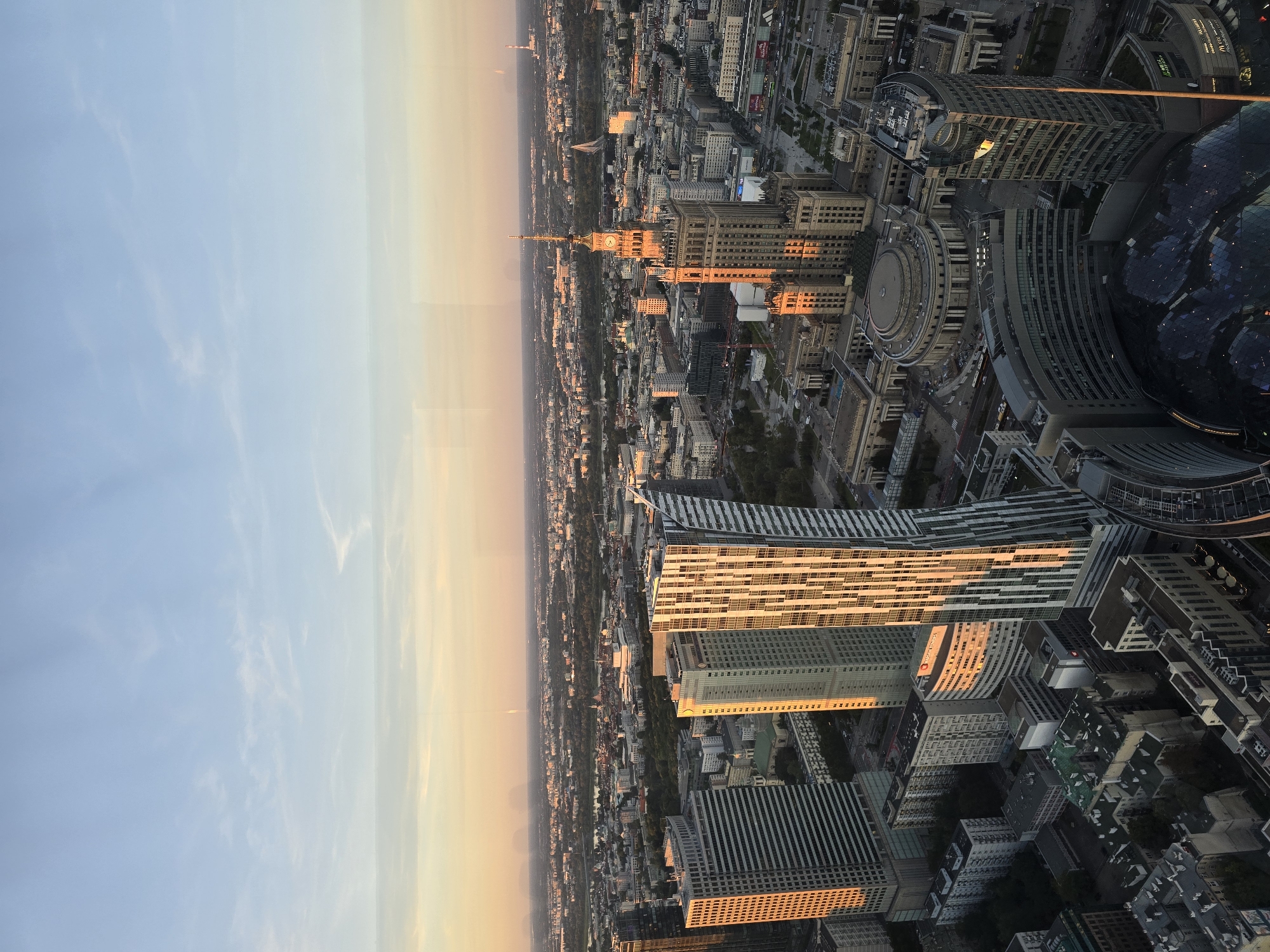 This is an aerial, high-angle, wide shot of the city of Warsaw, Poland, taken at dusk. The sky is a pale blue with wispy clouds, transitioning to an orange hue near the horizon. The foreground is dominated by several modern skyscrapers. To the left is a rectangular, grey skyscraper with horizontal window lines. To the right of that is a lighter-colored skyscraper with a rounded corner. In the center right, a prominent Stalinist-era skyscraper, the Palace of Culture and Science, stands tall with a clock face near its spire. Its facade is lit by the setting sun, highlighting its architectural details. Several other buildings of varying heights and architectural styles are visible across the cityscape, including a circular building in the mid-ground and a curved, glass-domed structure in the lower right. The city stretches into the distance, with a network of roads and what appears to be a body of water reflecting the warm light of dusk. There are no visible people or vehicles in the frame. The overall impression is a panoramic view of a dense urban landscape bathed in the warm, fading light of sunset.