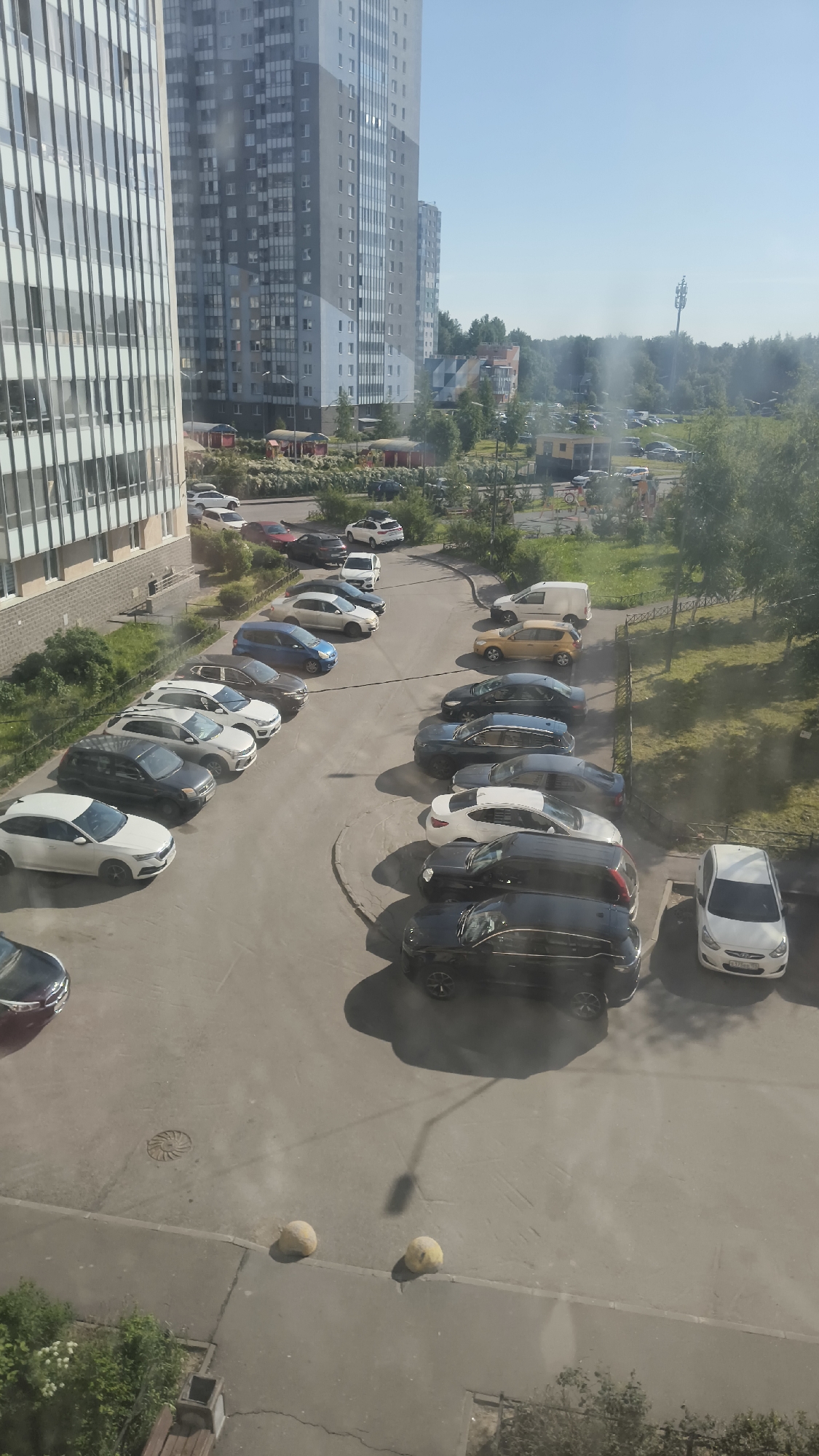 An overhead view captures a residential parking lot surrounded by modern multi-story buildings and green spaces on a bright, sunny day. The scene depicts a dense urban or suburban environment with numerous parked cars.
On the left, a tall building with a facade featuring many vertical window strips stands prominently. To its right and in the background, another large apartment building exhibits a gray and light blue checkered pattern on its exterior. Several smaller buildings and lush trees are visible in the middle distance, along with a cellular tower against the clear blue sky.
The main subject is a large asphalt parking area filled with various models and colors of cars, including sedans, SUVs, and vans. The vehicles are parked in rows and along curved paths that crisscross the lot. Notable car colors include white, black, gray, blue, red, and a yellow/gold sedan. Shadows are cast by the cars and buildings, indicating a sunny midday or early afternoon. No people are visible, and the primary activity is parking.
In the foreground, two yellow, spherical bollards sit on the edge of a paved area. A manhole cover is also visible on the asphalt. To the far right, a white Hyundai Creta SUV is parked; its license plate reads "A158MM 178," with "178" being a Russian region code, suggesting the location is likely St. Petersburg, Russia.
