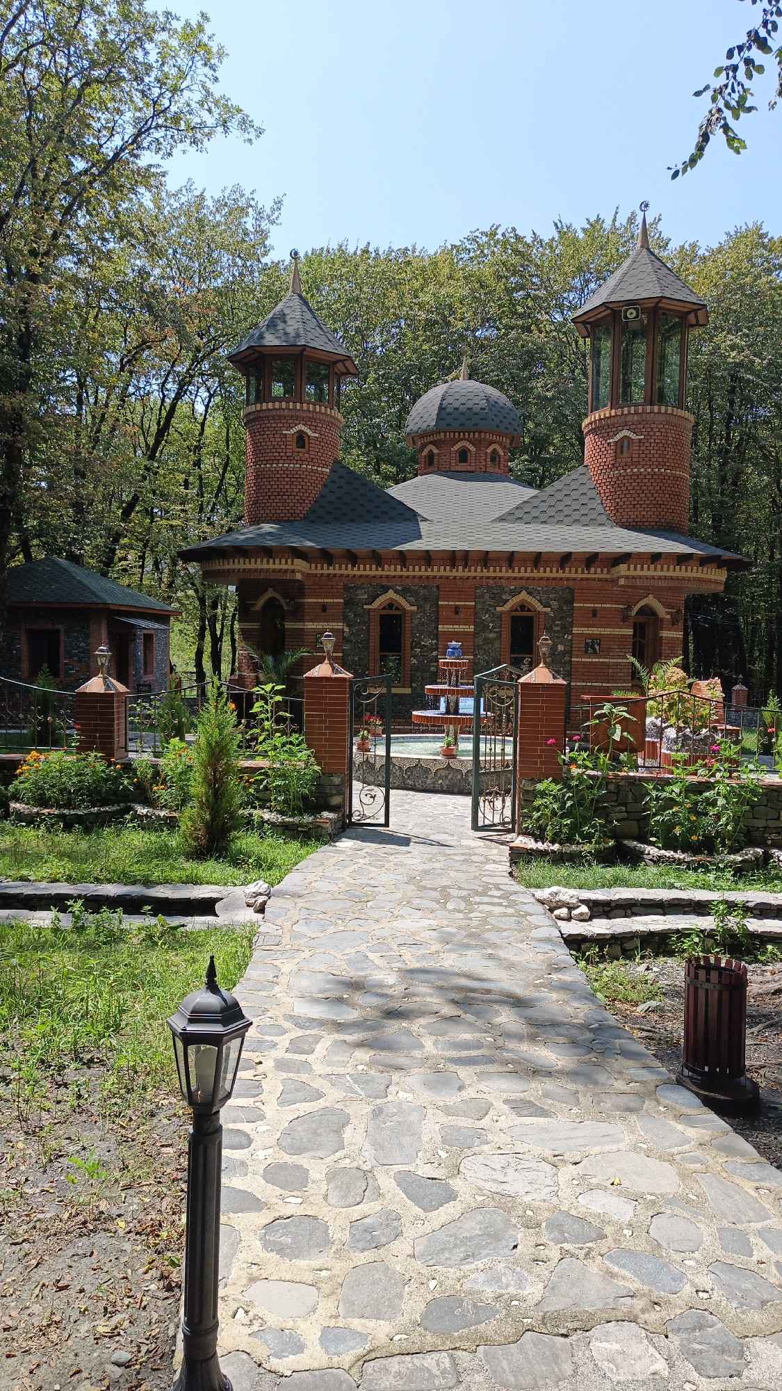 This is a full-shot, eye-level, outdoor photograph of the exterior of a mosque with two minarets and a dome, located in the city of Gabala, Azerbaijan. The mosque's facade is constructed from red brick and dark stone, with arched windows and decorative elements. In front of the mosque, there is a stone pathway leading towards an open gate. Beyond the gate, a small fountain with cascading water is visible. The area surrounding the mosque is landscaped with green grass, flowering plants, and trees. A black lamppost stands to the left of the pathway, and a wooden trash bin is located to the right. The sky above is clear and blue.