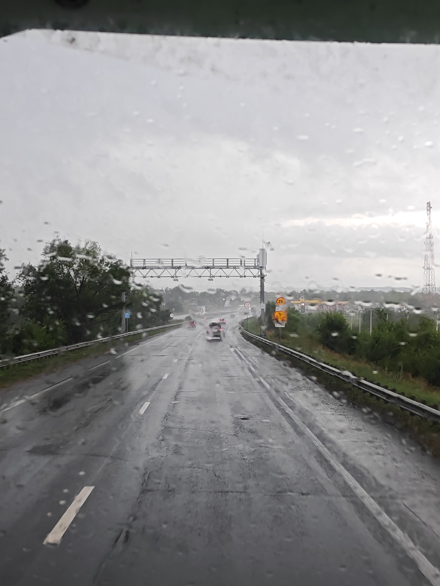The image captures a view from inside a vehicle driving on a wet highway under an overcast sky. Raindrops are visible on the windshield, distorting the view of the road ahead. The highway appears to be multi-lane, with vehicles driving in the distance. Green trees and vegetation line the sides of the road, and a guardrail is visible. Several road signs are present, including a yellow circular sign with the number "25" inside, and a sign indicating a speed limit of "70". In the background, there are buildings that appear to be part of a town or service area, including a gas station with yellow canopies. A tall telecommunications tower is also visible in the distance. The overall atmosphere is gloomy due to the rainy weather. The scene is consistent with a highway in or near Belozerki, Russia.