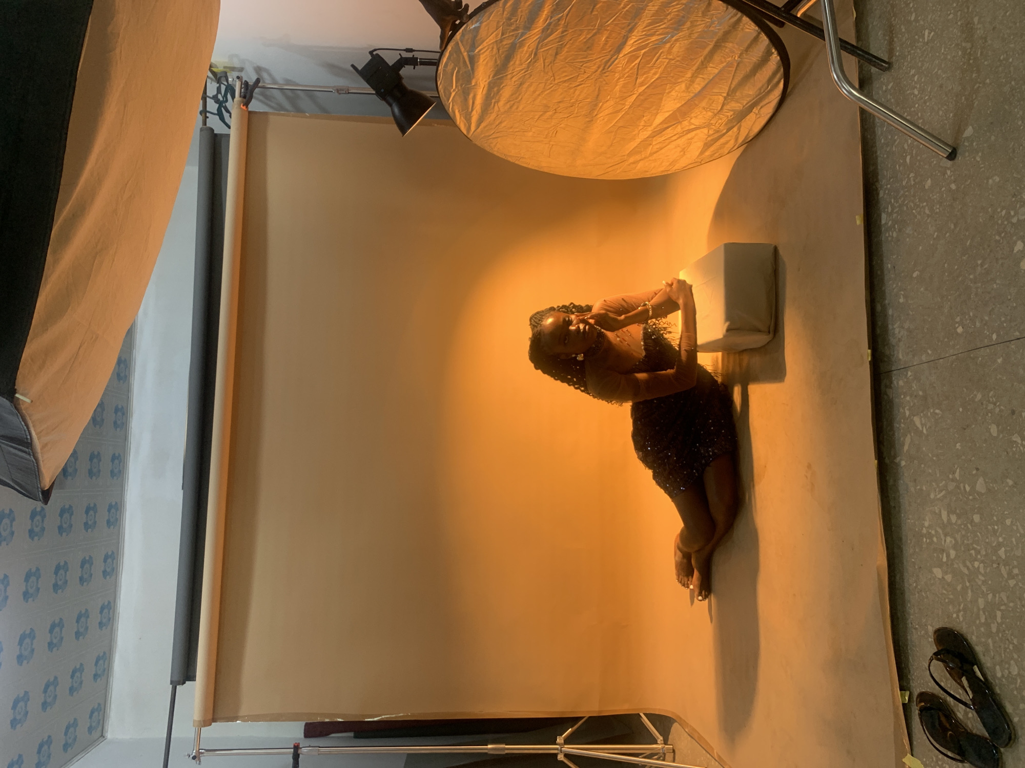 A young black woman is kneeling on the floor in a photography studio with a tan backdrop, with her hands clasped together near a white padded cube. She has braids in her hair and is wearing a dark, sequined dress. She is lit by studio lighting, including a large reflector above her and a softbox to her left. The floor is concrete with speckled gray stones and a pair of black flip-flops are located in the bottom right corner. In the background, there are studio equipment stands and rollers for the backdrop. The location appears to be an indoor studio space located in Gura Top, Nigeria.