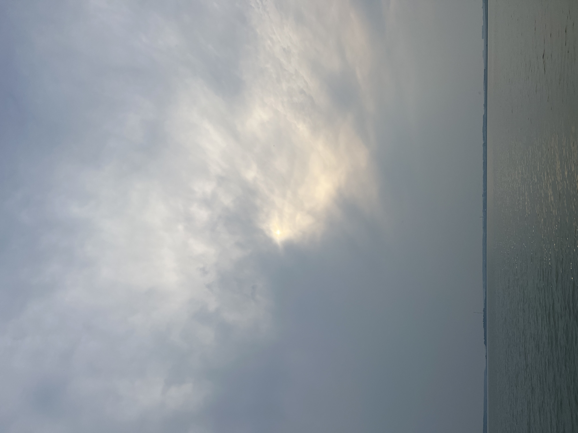 The image shows a wide expanse of water with a hazy horizon. The sky is overcast with scattered clouds, and the sun is partially visible, casting a soft glow. In the distance, a faint line of land is visible with what appears to be buildings and a tall structure, possibly a radio tower, on the horizon. The water is calm with gentle ripples. The scene suggests a natural landscape, likely a body of water such as a river or lake, with hints of civilization on the opposite shore. The location in Novopichugovo, Russia is not directly evident from the visual cues within the image itself, but the description implies this as the setting. There are no people or specific actions depicted in the image.