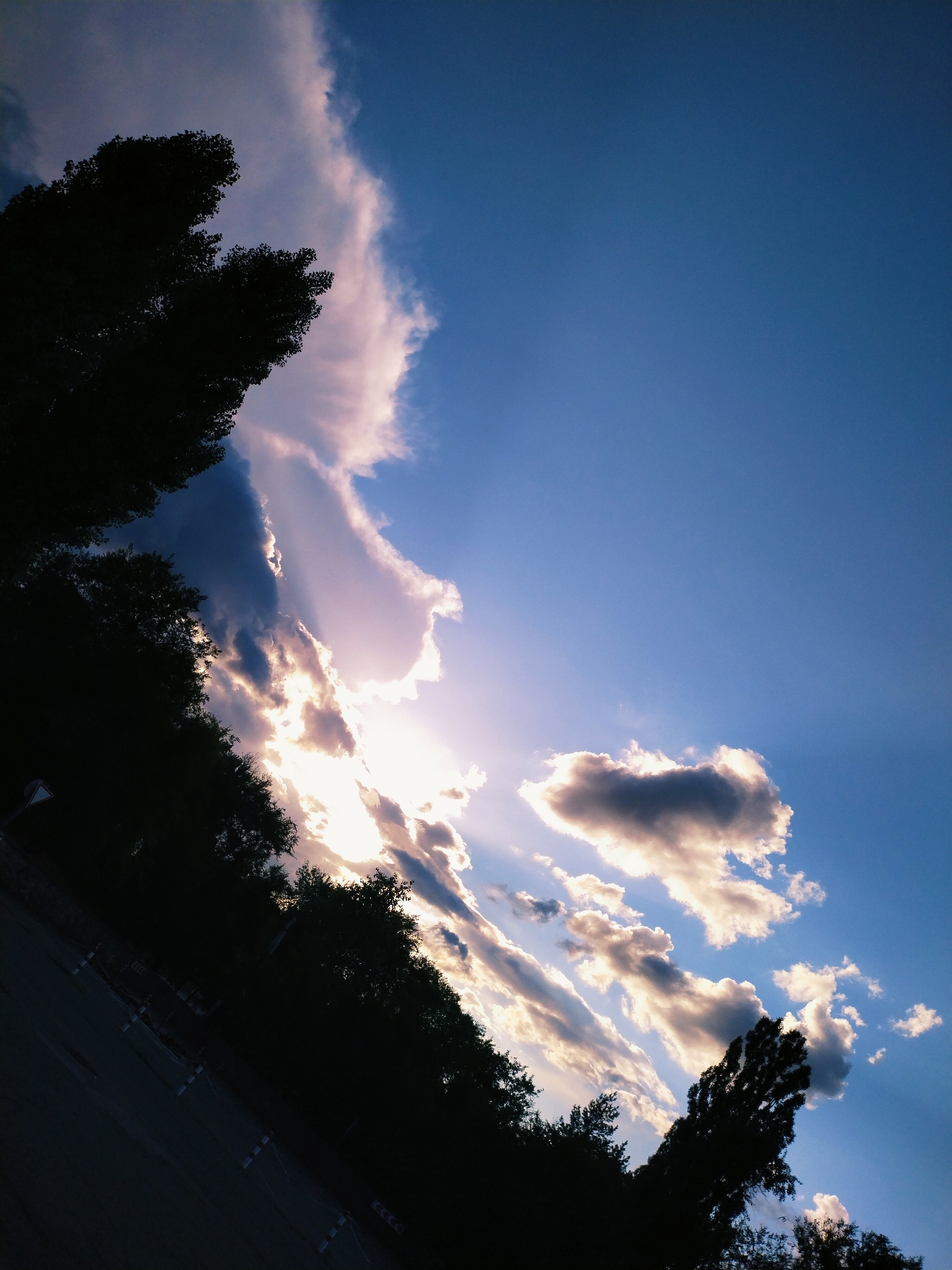 The image depicts a view of the sky with dramatic clouds, illuminated by the sun, seen from a silhouetted tree-lined area. The perspective is angled, suggesting the photo was taken from a low vantage point looking upwards. The environment appears to be an outdoor setting, potentially a park or roadside, with a dark foreground that includes a road and some traffic cones. The scene is likely in Horishni Plavni, Ukraine, given the context. There are no people or discernable actions visible in the image.