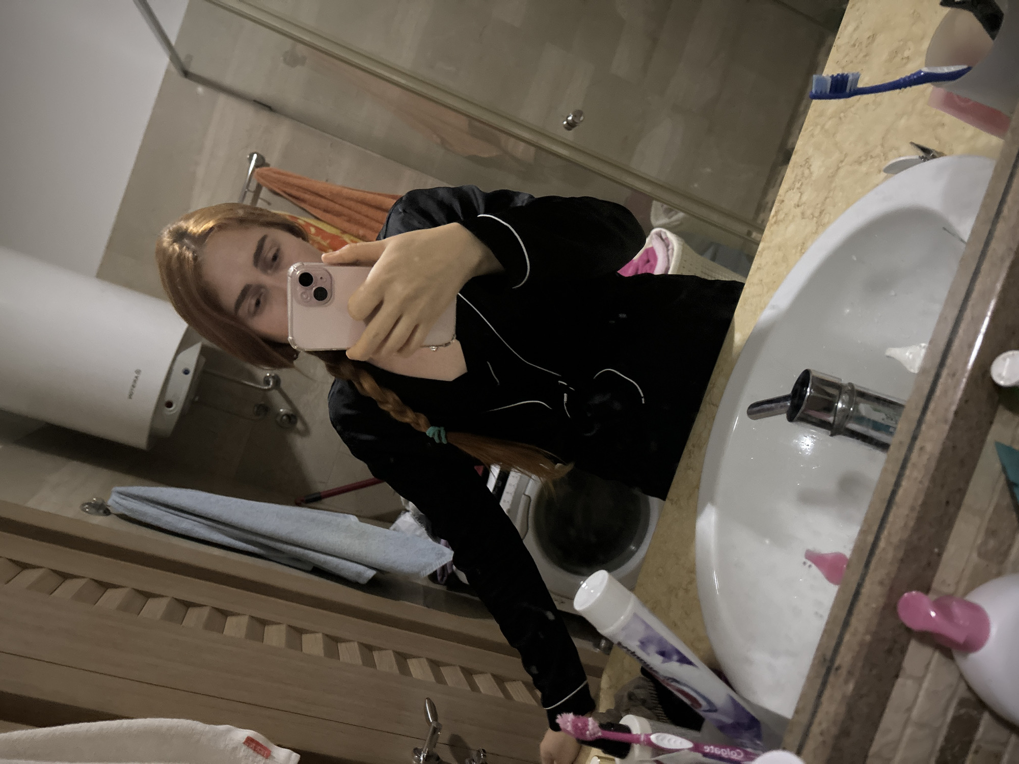 A woman with reddish-blonde hair is taking a selfie in a bathroom. She is wearing black pajamas with white trim. She is holding a pink smartphone. The bathroom contains a sink, a mirror, various toiletries, a water heater, and a washing machine. The location is most likely a private residence in Budva, Montenegro.