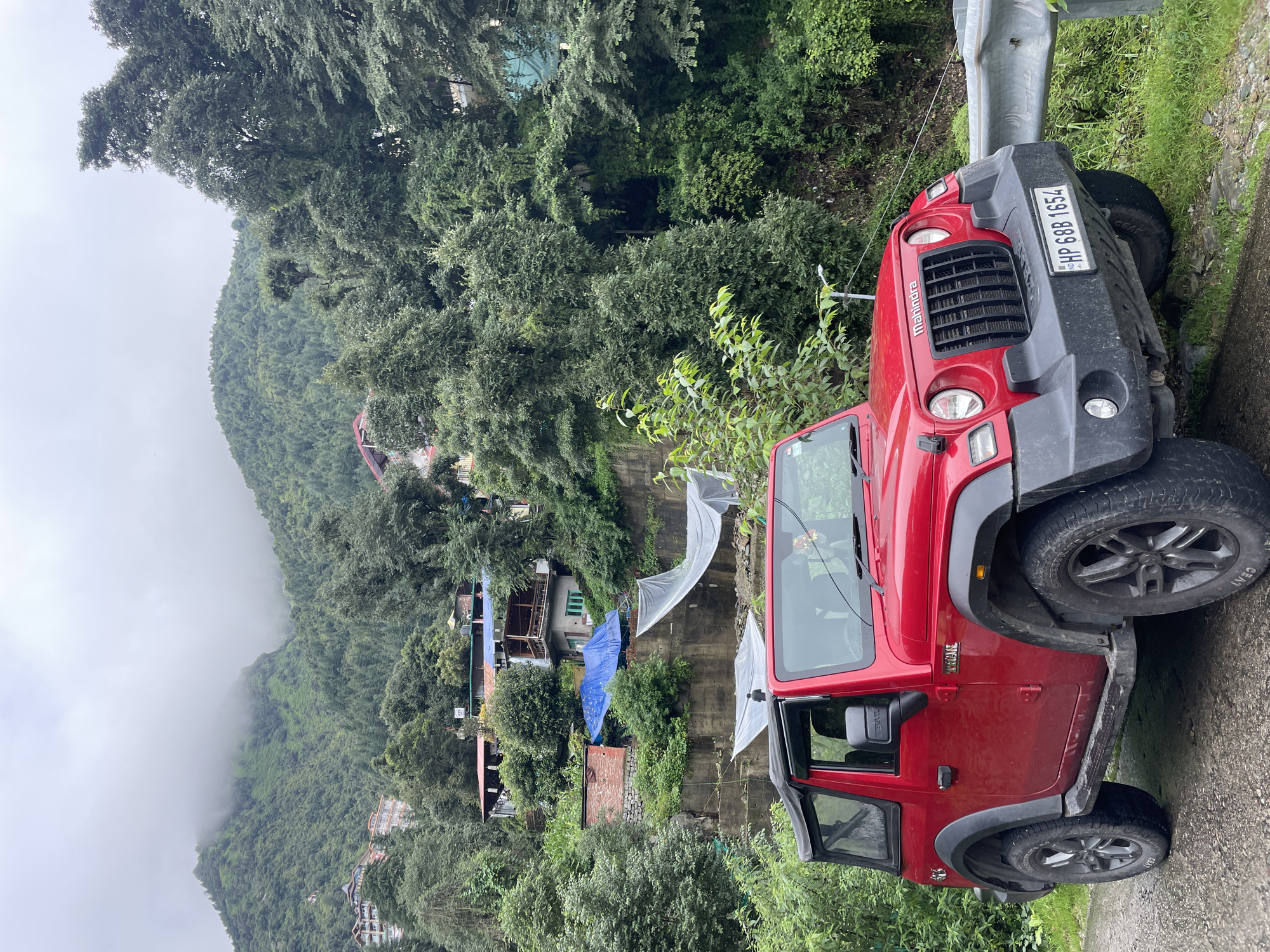 A bright red Mahindra Thar is parked on the side of a narrow, paved road in a hilly, forested area. The vehicle is facing slightly to the right. The road is a rough gravel and dirt surface. Beyond the road, a steep embankment rises, covered in lush green trees and vegetation.
In the background, several buildings are visible, nestled into the hillside. These appear to be houses or small guesthouses with red-tiled roofs and balconies. Some structures are covered with blue and white tarpaulins, possibly for protection or construction. A concrete retaining wall is visible supporting the slope.
The terrain is mountainous, with dense evergreen trees covering the slopes. The sky is overcast and a thick mist or fog is obscuring the upper reaches of the mountains. The overall atmosphere suggests a damp, cloudy day in a scenic, elevated location. A license plate on the front of the car reads "HP 688 1654".