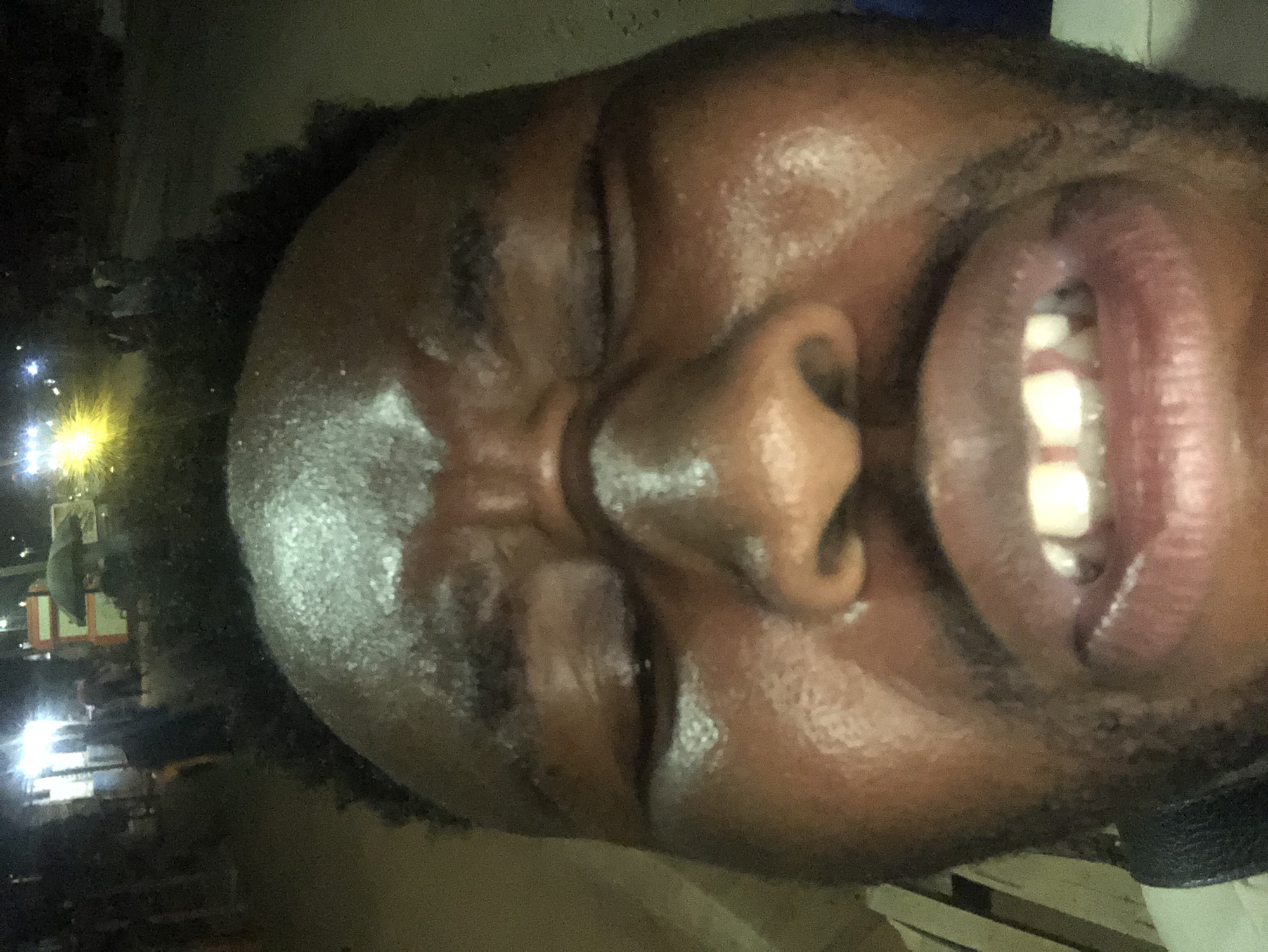 A close-up, slightly angled photograph shows the face of a Black male with dark, curly hair. His eyes are closed, and his mouth is open, revealing his teeth. The skin appears slightly glossy. The background is dark and indistinct, but some lights and the vague shapes of buildings suggest an urban environment at night. The city and state are not specified in the image.