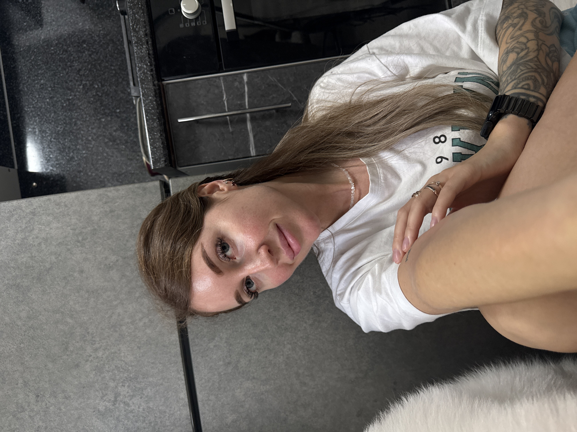 A medium shot photograph shows a woman with brown hair and blue eyes wearing a white t-shirt. She is sitting down, with her right arm resting on her leg and her left arm on her chest. Her right hand is on her left arm, and she is wearing rings on her fingers. She has a watch on her left wrist. The background is a kitchen with dark countertops and cabinets. A gray wall is visible on the left side of the image. The woman is looking at the camera and smiling. The photograph is taken indoors. The city of Chaikovsky, Russia is not identifiable from the image.