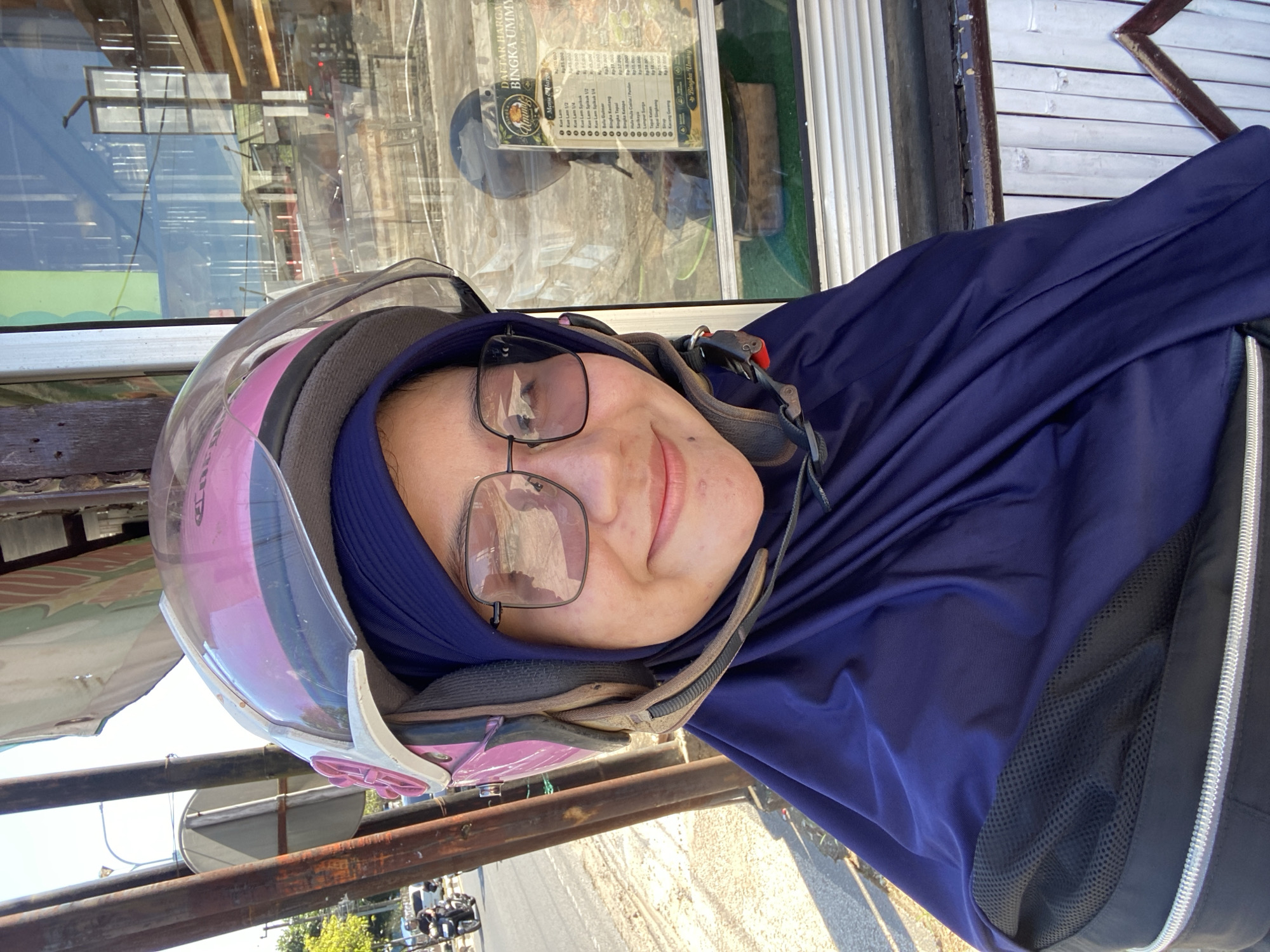 A woman wearing a dark blue hijab and a pink helmet with a clear visor is smiling at the camera. She is also wearing square-shaped glasses. Her face is partially obscured by the helmet and hijab. The woman appears to be standing outdoors, possibly on a street, as evidenced by the background which includes a road with a motorcycle in the distance and some vegetation. To her right, there is a glass window reflecting buildings and possibly the sky. A price list is visible on the window, suggesting the location might be a food stall or shop. The overall scene is brightly lit by sunlight. The location is identified as Sungai Paring, Indonesia.