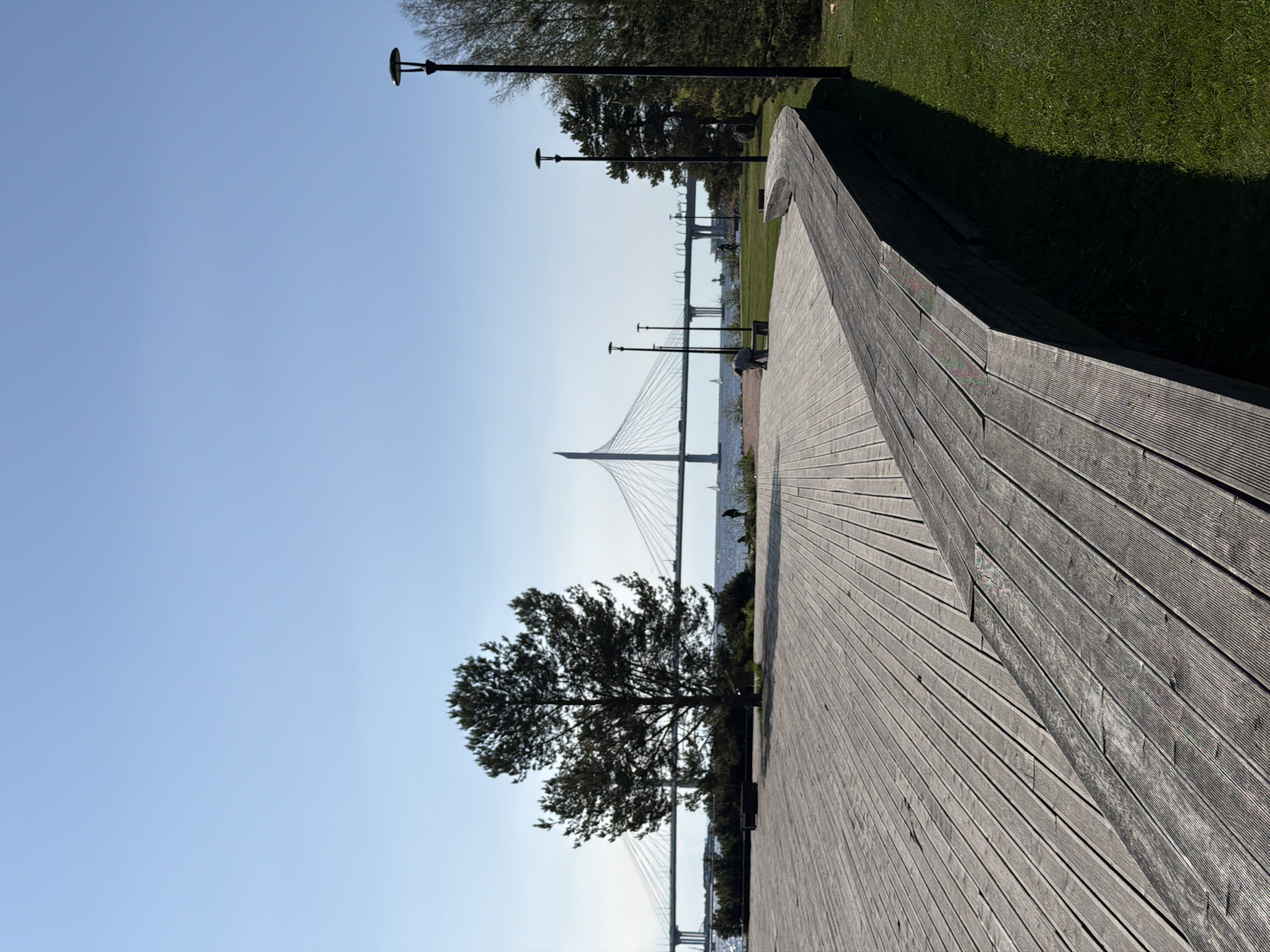 The image captures a view along a wooden boardwalk or deck, possibly a recreational area or park. The decking runs diagonally across the frame, its planks showing distinct grain and texture. Adjacent to the boardwalk is a patch of green grass, indicating a park or landscaped area.
In the background, a modern cable-stayed bridge spans a body of water. The bridge is a significant architectural element, with tall pylons supporting a network of cables. In the distance, additional lampposts or lighting fixtures are visible, suggesting an urban setting. A few people can be seen in the distance, though they are not the primary focus of the image. A tree is growing along the boardwalk, partially obscuring the view of the bridge.
Based on the bridge's architectural style and location, it can be inferred that the image was taken in Saint Petersburg, Russia, with the bridge likely being the Western High-Speed Diameter (WHSD) bridge.