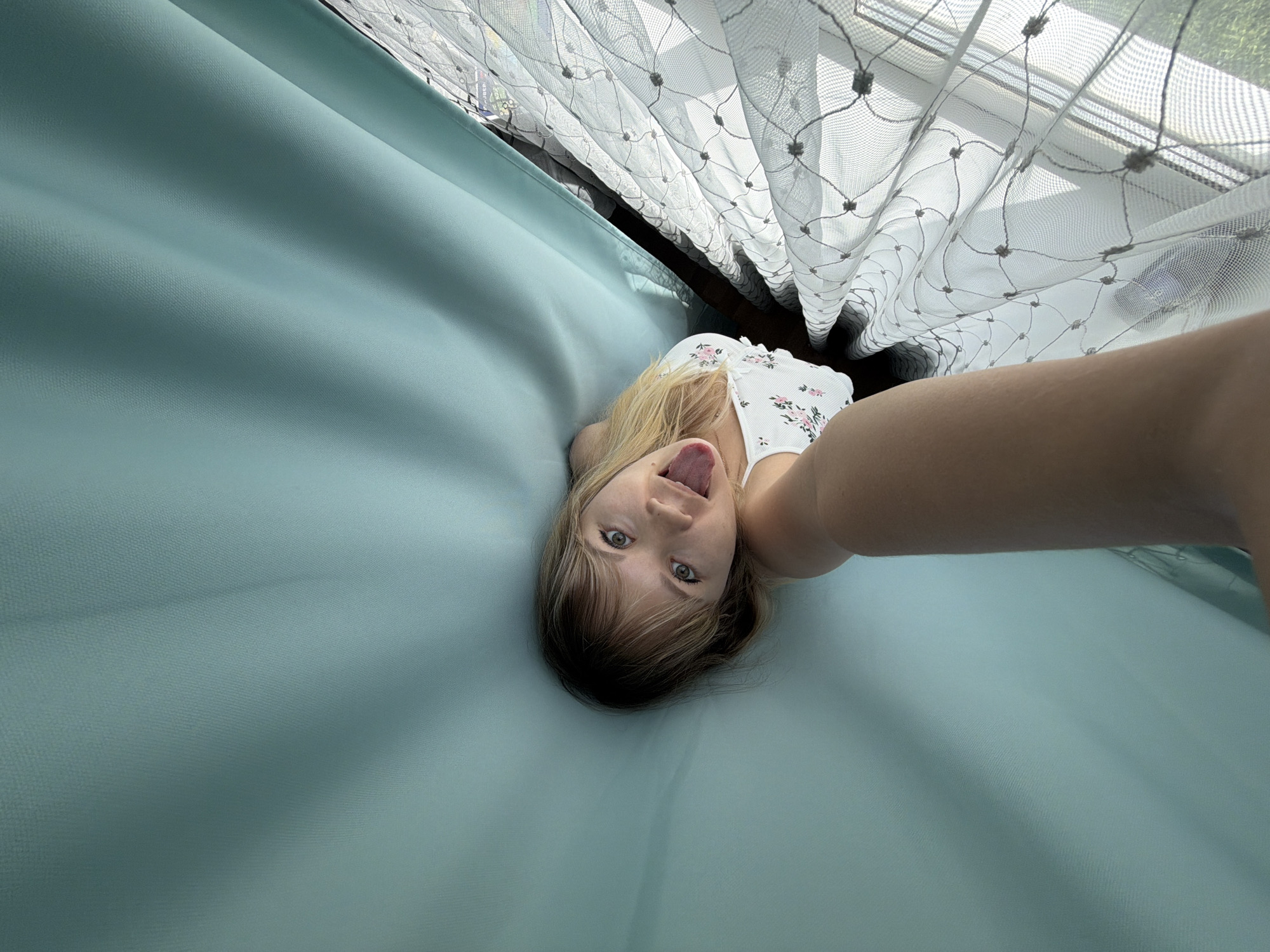 A woman with blonde hair is lying on a light blue sheet, with her face turned towards the camera and her tongue sticking out. She is wearing a white top with pink flower patterns. Above her, there is a lace curtain partially visible, and beyond that, a window. The photo appears to be taken indoors, possibly in a bedroom. There is no specific information to pinpoint the location as Chaikovsky, Russia.