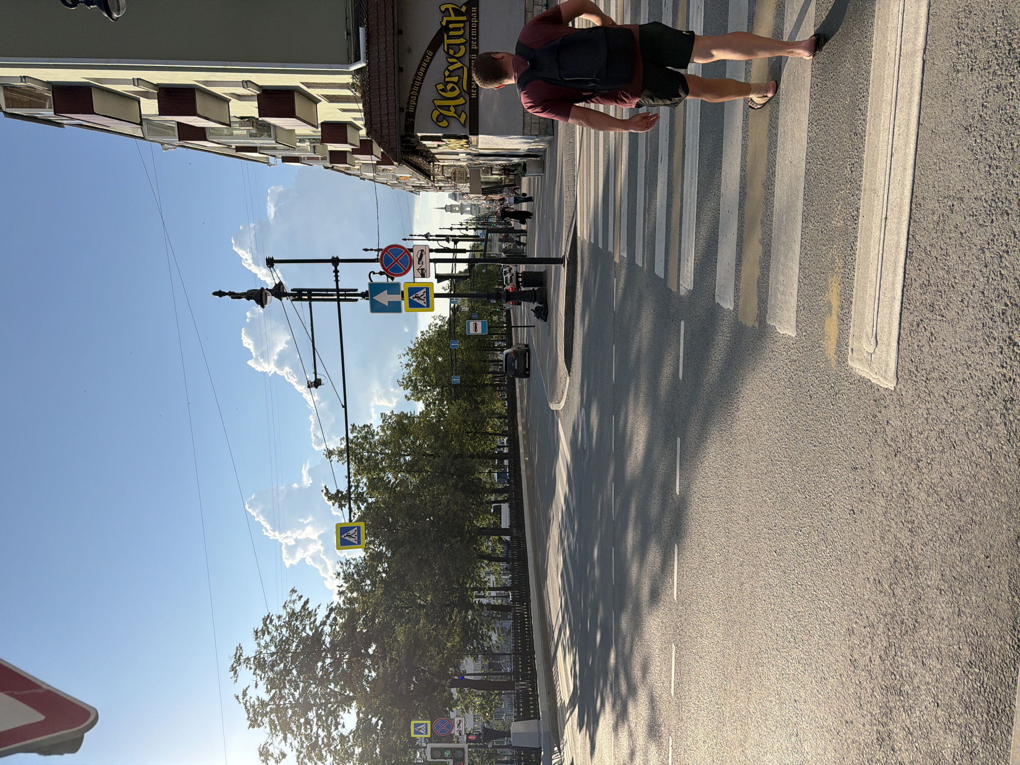 The image depicts a street scene in Perm, Russia, likely on a sunny day. In the foreground, a pedestrian with a backpack is crossing a marked pedestrian crossing on a paved road. The pedestrian is wearing a maroon t-shirt and black shorts. To the right of the pedestrian, a building with several balconies is visible. The street is lined with trees, and in the distance, architectural elements like towers are present, suggesting a city environment. Several traffic signs and streetlights are also visible along the road, indicating this is an urban area. The overall atmosphere is that of a typical day in a Russian city, with elements of daily life and infrastructure.