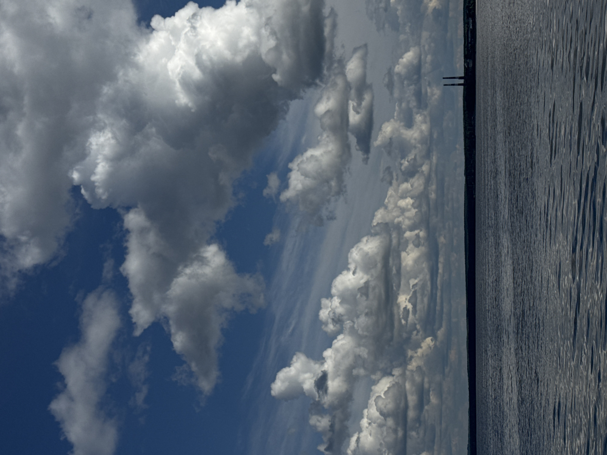 The image shows a body of water adjacent to a land mass under a sky with cumulus clouds. The water surface is rippled, suggesting wind or current. A dark line separates the water from the land, possibly indicating a shoreline with vegetation. Two vertical, pole-like structures are visible on the landmass, possibly pilings or chimneys, providing a sense of scale. The clouds are a mix of white and gray, contrasting against the blue sky. The location is Chaikovsky, Russia.