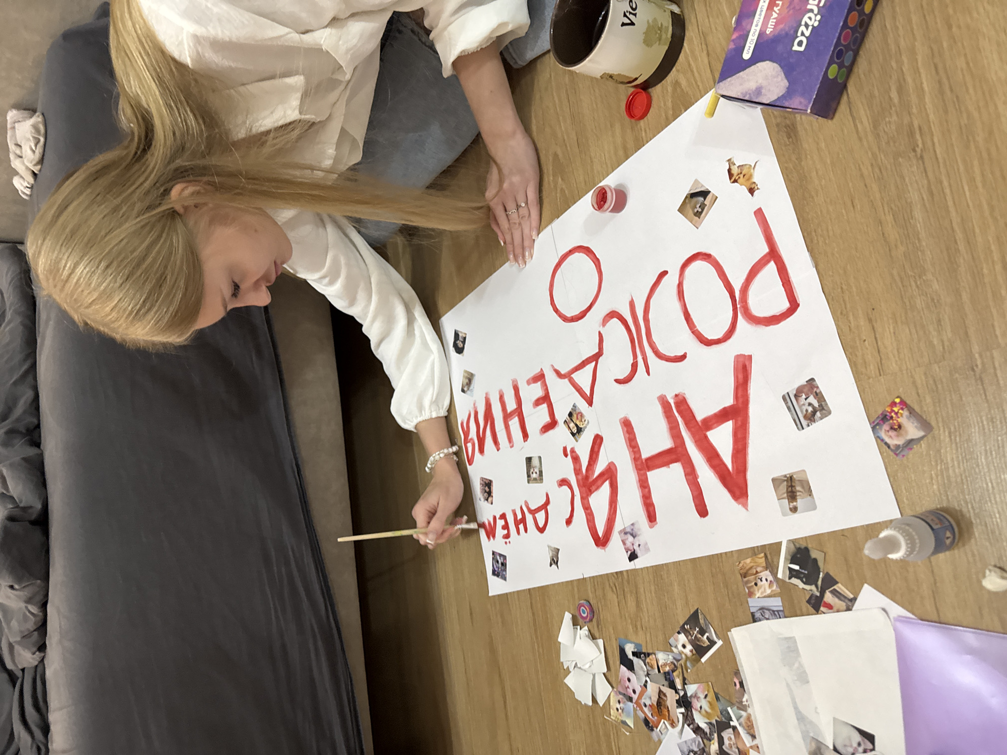 A young woman with long blonde hair sits on the floor, focused on a large white poster. She is wearing a white shirt and blue jeans and is holding a paintbrush in her right hand, adding red paint to the poster. The poster has large red Cyrillic letters that spell out "С ДНЁМ РОЖДЕНИЯ" (Happy Birthday). Small photos of what appear to be cats are glued around the text.
Various art supplies are scattered around the poster, including a small container of red paint, a red paint cap, a pencil, and a box of colored pencils or crayons labeled "Grëza". A metal can with writing on it is also nearby. The setting appears to be a living room, with a grey couch visible to the left of the frame and a wooden floor. The lighting suggests it's daytime. The woman's focused expression and the creative activity indicate she is preparing a handmade birthday greeting.