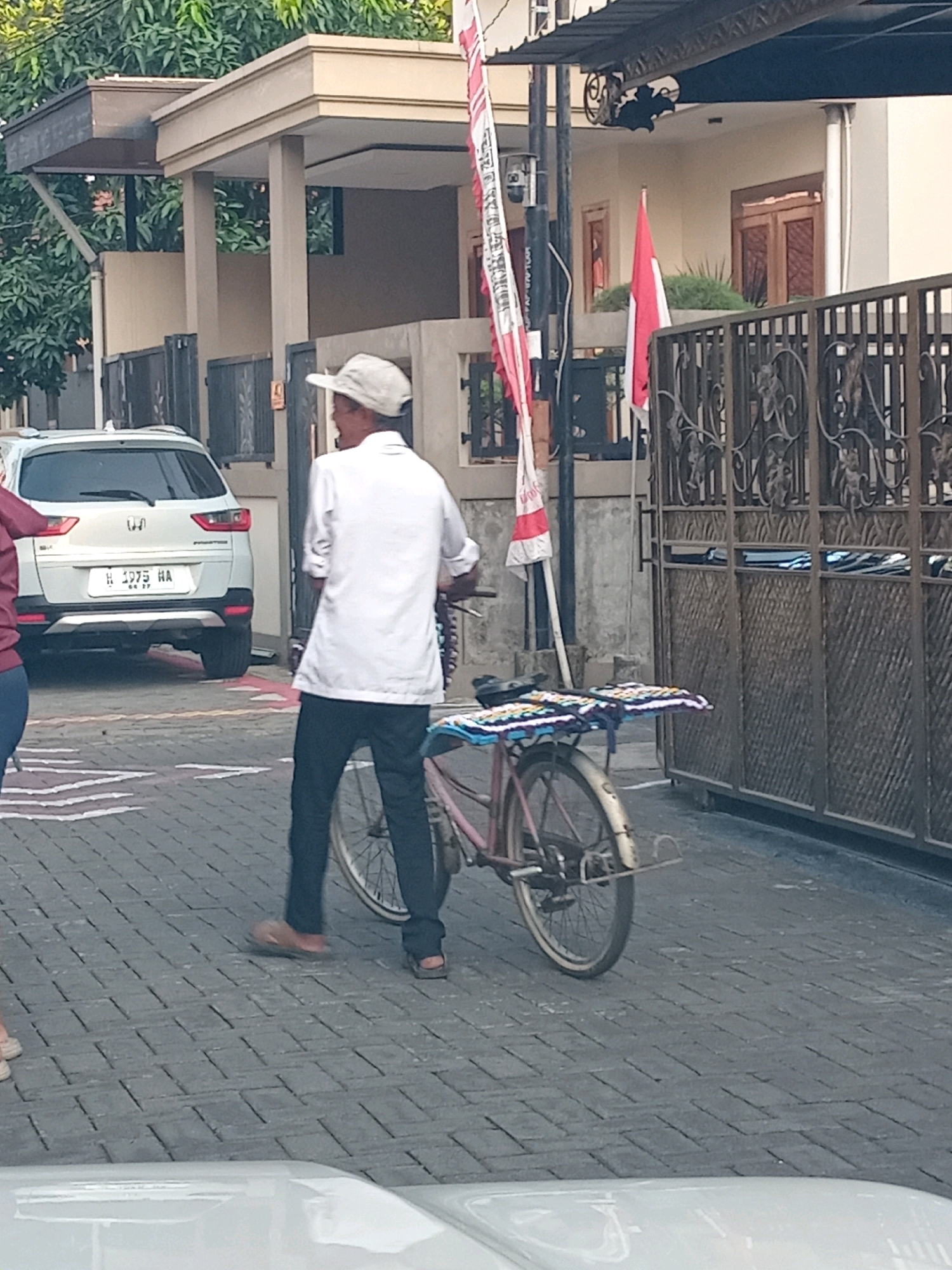 The image depicts a street scene in Semarang, Indonesia. In the foreground, a man wearing a white cap and a white shirt, with his sleeves rolled up, stands next to a pink bicycle. He is wearing dark pants and sandals. The bicycle has a rack on the back with two colorful woven mats. To the left of the man and bicycle, a white SUV is parked, with its license plate reading "H 1975 HA". Behind the man and bicycle, a tall metal gate with ornate scrollwork is visible. Further in the background, a building with a tiled roof and wooden window frames is present. A red and white Indonesian flag is displayed on a pole to the left of the gate. The ground is paved with interlocking bricks. Trees are visible in the upper left corner of the frame. The scene appears to be a residential street.