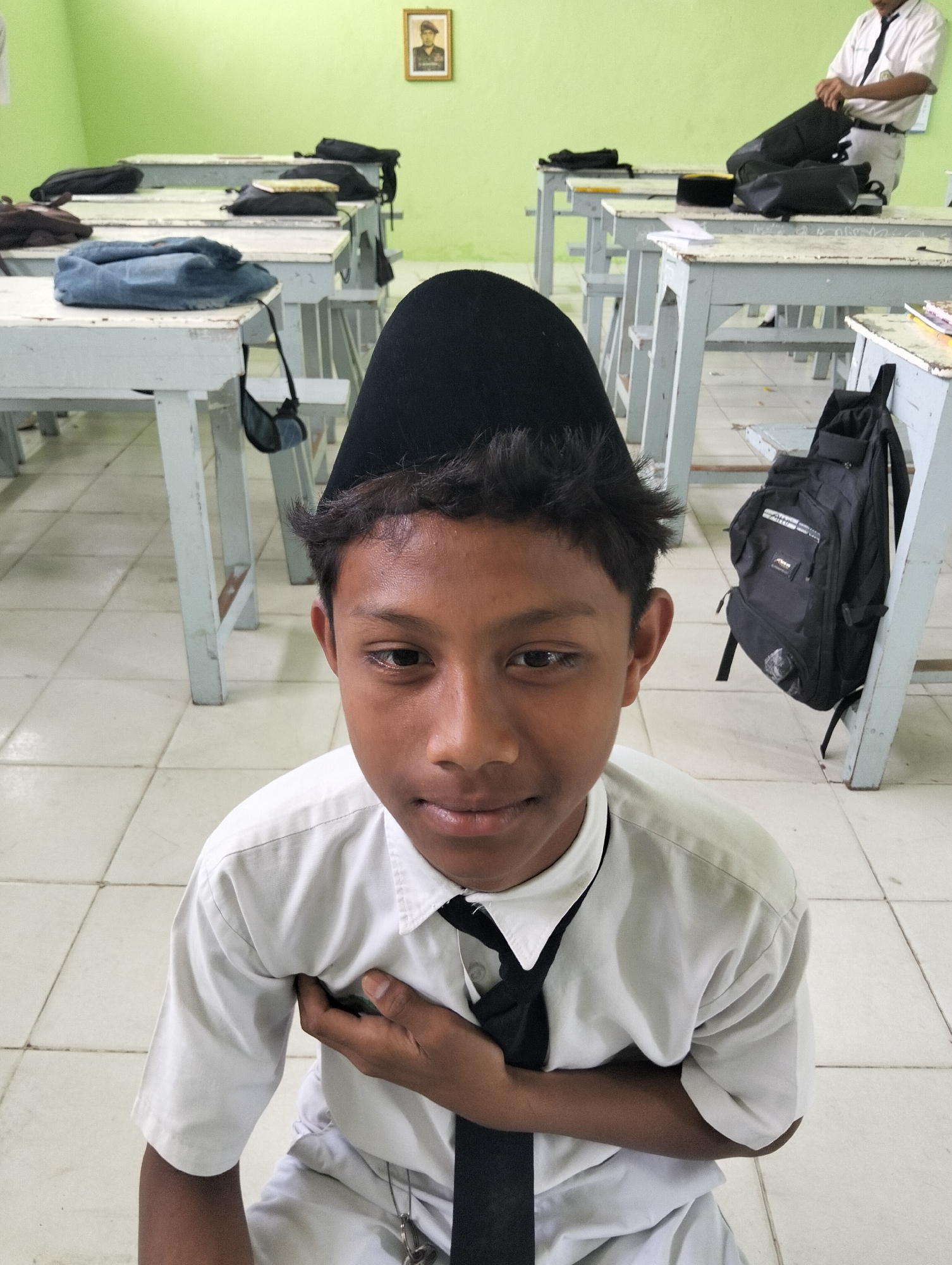 A young boy wearing a white school uniform, including a black tie and a black kufi cap, is seated on a tiled floor in a classroom. He is looking directly at the camera with his hand placed on his chest. Behind him, several rows of wooden desks with white tops are visible. Backpacks are placed on some of the desks and on the floor. In the background, a framed portrait of a person in uniform hangs on a pale green wall. The overall environment suggests a school setting in Kranji, Indonesia.