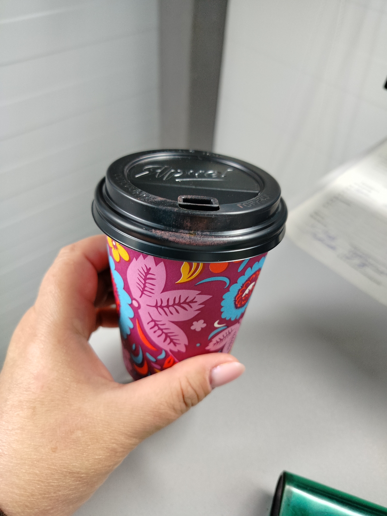 A hand holds a disposable coffee cup with a black lid. The cup has a colorful floral pattern. The lid is slightly ajar and appears to have some reddish residue around the opening. In the background, there is a white, horizontally lined wall and a desk with some documents. The scene is indoors. The city of Yashkino, Russia is not depicted in this image.