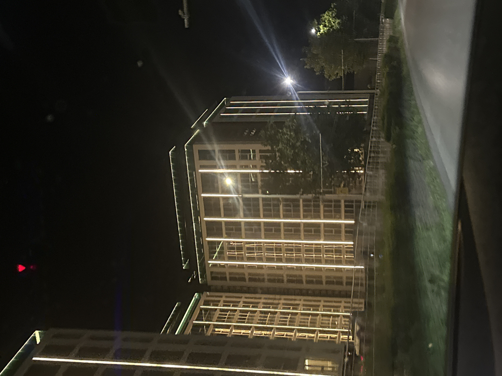 The image is a night-time photograph taken from a moving vehicle, likely on a road. In the background, there are two tall, modern apartment buildings with illuminated vertical lines of light running along their facades. The buildings appear to be under construction or newly built, as suggested by the minimalistic lighting design. Green lights are visible along the top edges of the buildings. Trees and vegetation are present in the mid-ground, with some of them also illuminated by streetlights. A road is visible in the foreground, suggesting the vantage point of the photographer. In the sky, a red triangular light is visible, possibly an aviation warning light. The overall scene depicts an urban environment at night. The buildings are located in the city of Ruse, Bulgaria, specifically in the area of a new residential complex near the Danube River.