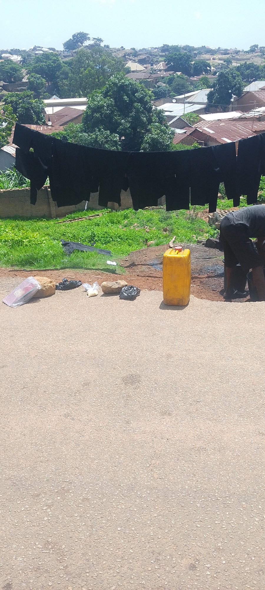 The image depicts an outdoor scene in Jos, Nigeria, under a clear, bright sky, suggesting daytime. The foreground is dominated by a paved road with a rough, grey asphalt surface. Scattered on the edge of the road are several items: a transparent plastic bag containing what appears to be white powdery substance, two round, brown rocks, two black plastic bags filled with unspecified contents, and a small plastic bag with white grains. To the right, a large, yellow plastic jerrycan stands upright.
Further back, a person wearing dark clothing is visible, bent over and seemingly engaged in some activity near the jerrycan and what looks like dark liquid on the ground. Behind this immediate area, a lush green grassy slope is covered in dense vegetation.
Stretched across the scene, above the grassy slope, a clothesline is hung with numerous pieces of dark-colored fabric, likely laundry, drying in the open air. The fabric appears to be heavy, with the pieces hanging in long, straight lines.
In the background, a residential area is spread across a hilly landscape. The houses are characterized by their corrugated iron roofs, some appearing rusted, and a mixture of concrete and possibly mud-brick walls. Trees are interspersed among the buildings, and rocky outcrops are visible, indicating the terrain of the area. The overall impression is of a typical residential setting in a somewhat rural or semi-urban part of Jos, Nigeria, during daylight.