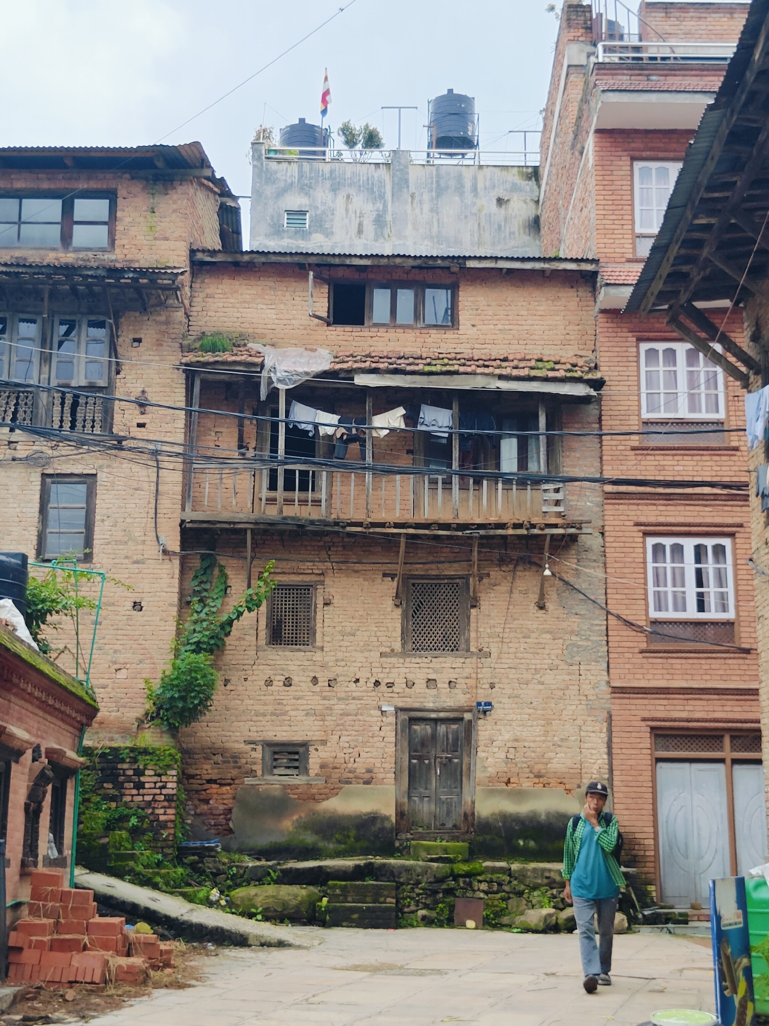 In Kirtipur, Nepal, a person walks on a cobbled street between two brick buildings. The person, wearing a plaid shirt, a turquoise t-shirt, jeans, and a baseball cap, carries a backpack and walks towards the right of the frame. The building on the left is older, with a weathered wooden balcony and laundry hanging out to dry. The building on the right is newer, made of red brick with white-framed windows and a modern doorway. The street is lined with greenery, including ivy growing on the older building and moss on the steps leading up to a doorway. The sky is overcast.