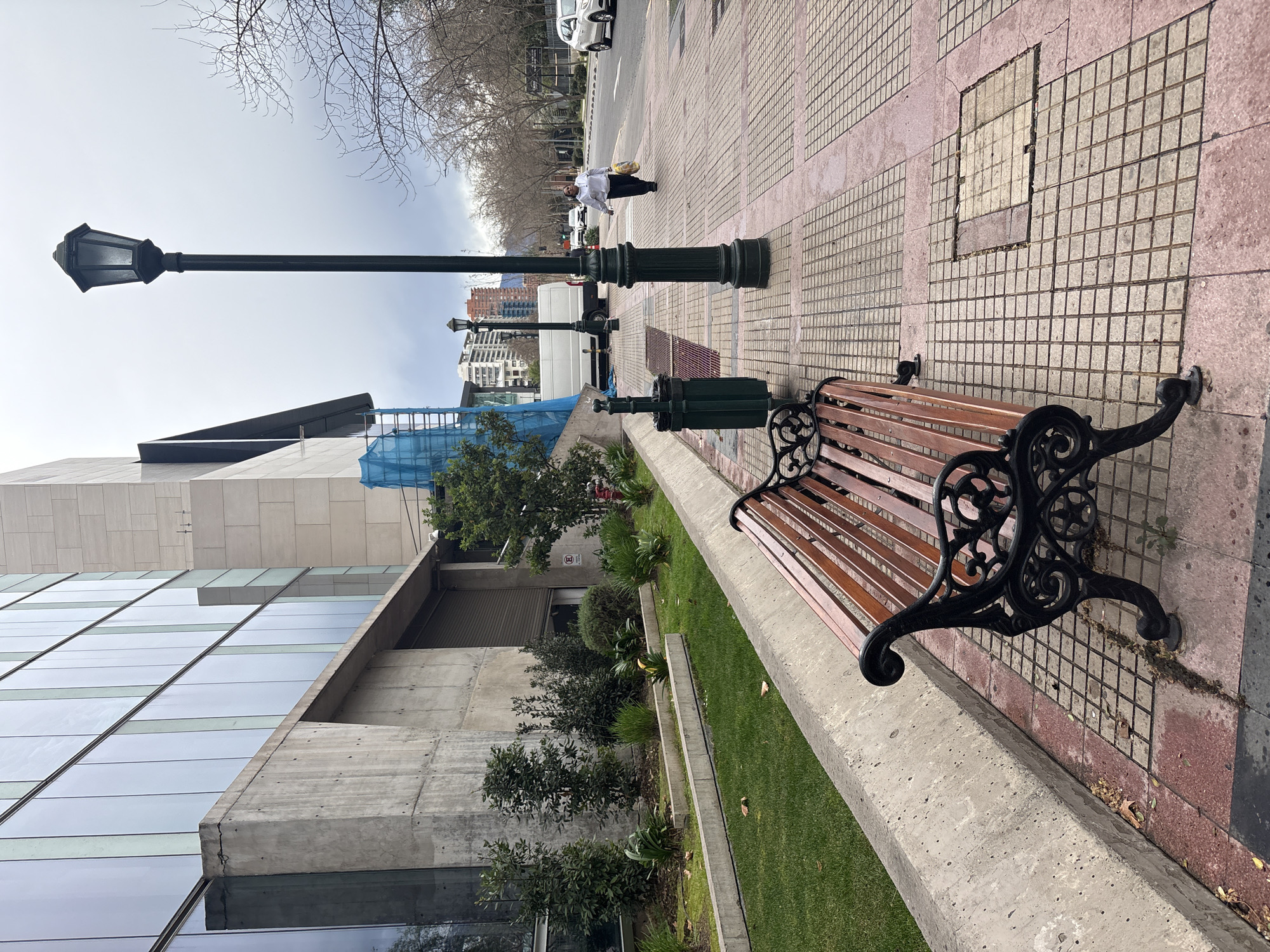 A full, eye-level shot shows a sidewalk with a park bench in the foreground, and a modern building and street scene in the background. The bench has a dark metal frame with wooden slats. To the left of the bench, there is a raised planter with grass and low-lying bushes. A dark green trash can is located near the bench. The sidewalk is made of red and beige square tiles, with some areas appearing wet. A black lamppost stands on the sidewalk, to the right of the bench. In the mid-ground, a woman wearing a white hooded sweatshirt and dark pants walks away from the camera, carrying a yellow bag. She is on the sidewalk, which runs alongside a street with moving vehicles. The street is lined with trees that have bare branches. In the background, there is a large, modern building with a glass facade and a concrete base. Construction scaffolding covered in blue sheeting is visible on the side of the building. Further in the background, more buildings and the sky, which is overcast, are visible. The scene is likely in Santiago, Chile, given the architectural style and vegetation.