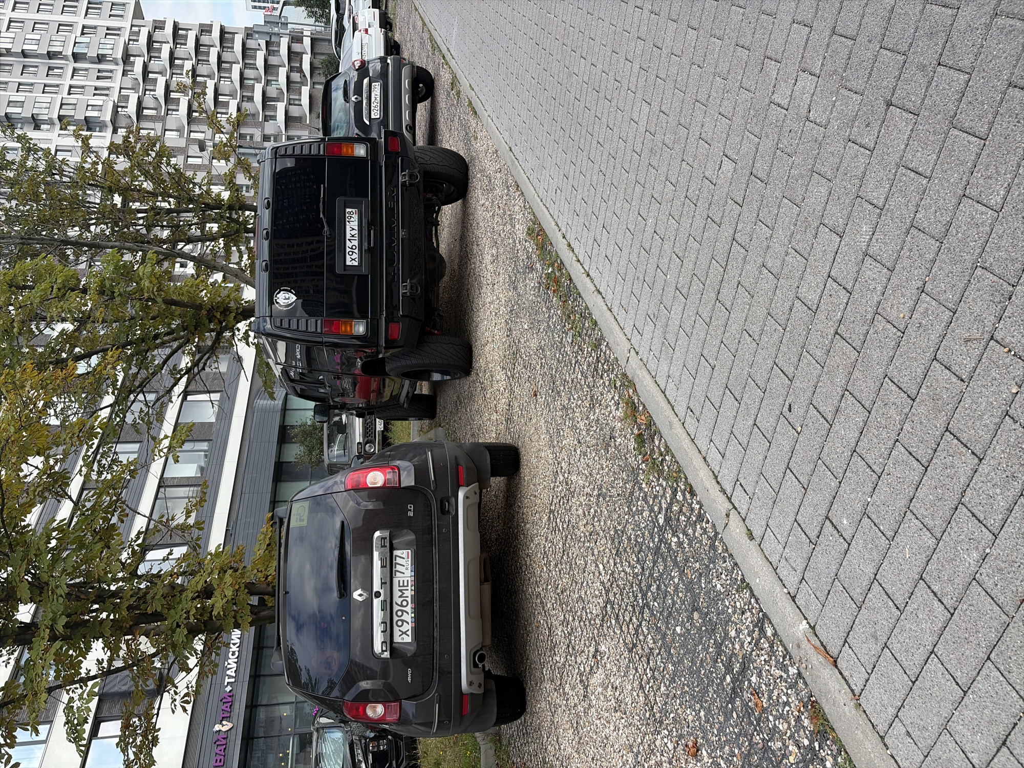 A black Hummer H2 SUV, license plate X961KY198, is parked next to a dark brown Renault Duster SUV, license plate X996ME777, on a gravel lot in front of a modern apartment building. The Hummer has a dark tint on its windows and a circular emblem on its rear door. The Duster has a roof rack and a spare tire on its rear door, and the license plate indicates it is a Russian vehicle. In the background, a multi-story residential building with balconies and large windows stands tall. The street is paved with gray interlocking bricks. Trees with green and yellow leaves are scattered throughout the scene, indicating an autumn setting. The overall environment appears to be an urban parking area.