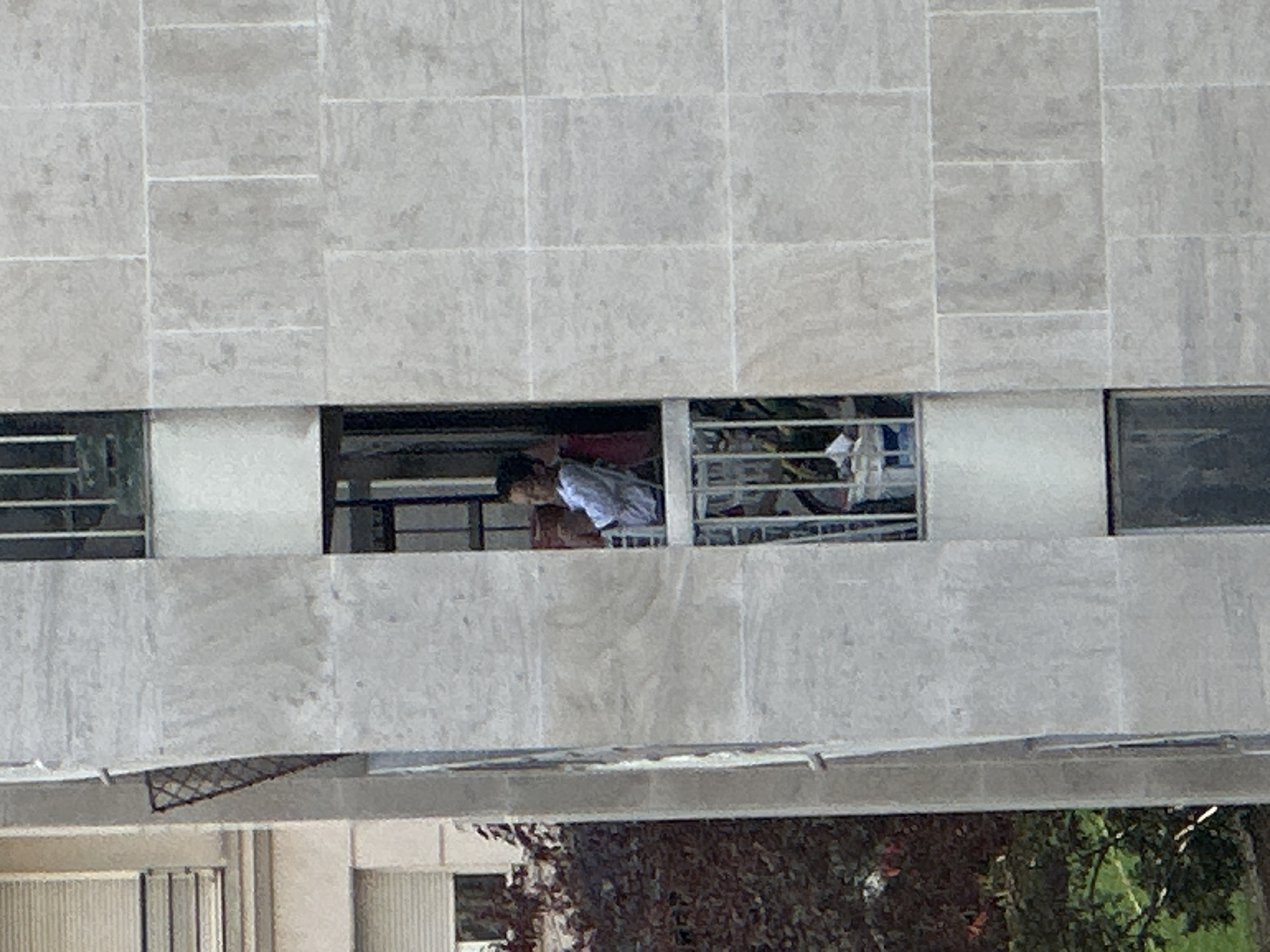 A person is visible on a balcony of a modern building, appearing to be in Paris, France. The individual, wearing a light-colored shirt, is seated and looking to their left. The balcony has a railing and a few objects are present, including what appears to be a red chair or cushion and some containers. The building's facade is constructed with large, light-colored stone tiles. To the left of the balcony, some dark foliage is visible. The scene is captured from a distance, looking up at the balcony.