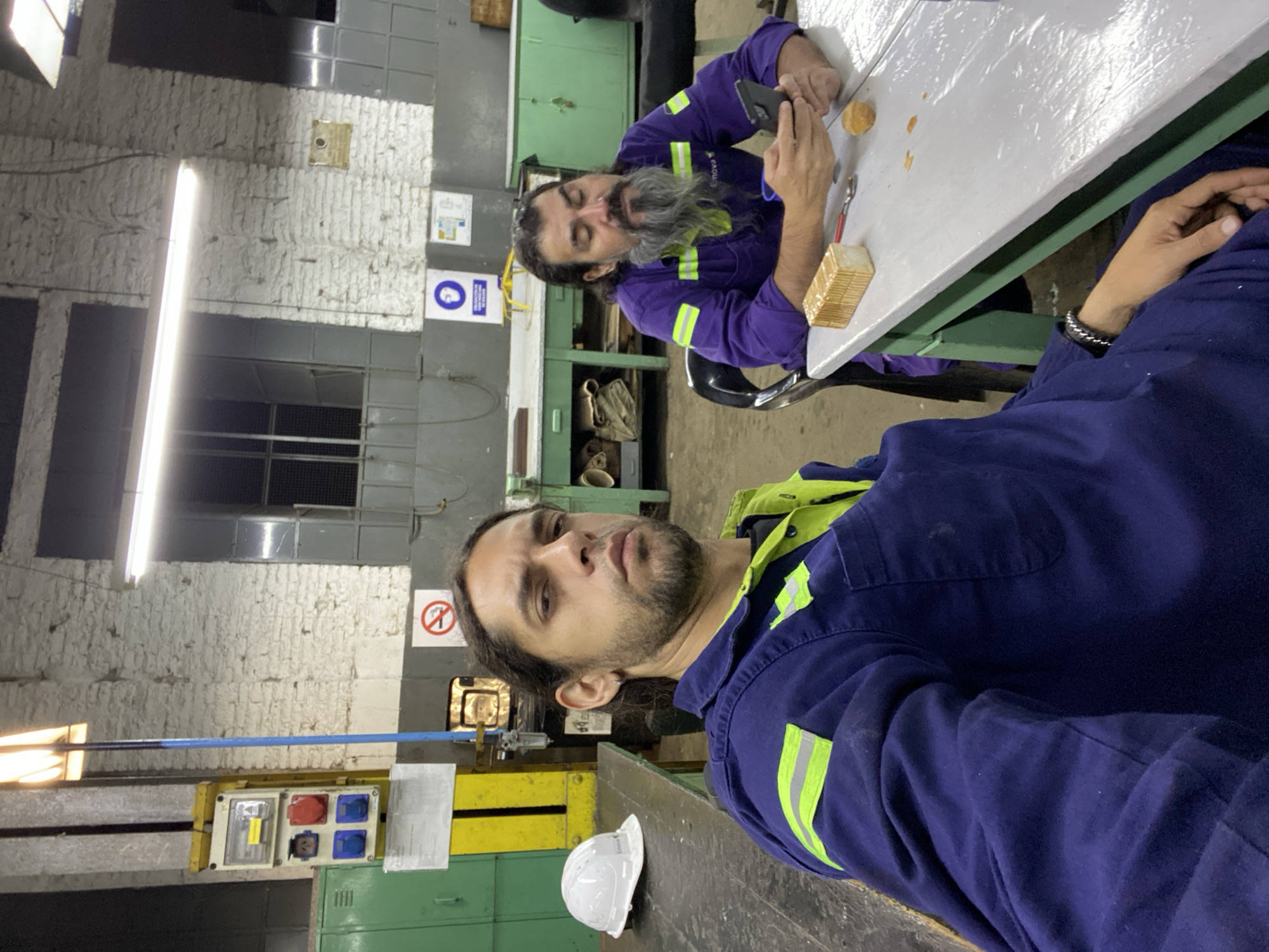 The image depicts an interior scene within an industrial or maintenance facility in Buenos Aires, Argentina. Two adult males are present in the foreground and midground. The man in the foreground, facing the camera with a direct gaze, has dark hair, a short beard, and an earring. He is wearing a navy blue work jumpsuit with neon green and silver reflective strips. To his left, on a dark workbench, rests a white hard hat with "Emova" branding.
In the midground, a second man with dark hair and a long grey beard is seated at a light grey table with a green metal frame. He is wearing a purple work jumpsuit with neon green and silver reflective strips, also bearing the "Emova" brand on his chest. He is looking down, holding and operating a black smartphone with both hands. On the table in front of him are a small loaf of white bread, a red-handled knife, a spoon, and a small food crumb.
The background features rough white-painted brick walls, metal-framed windows with grates, and various industrial elements. These include green metal lockers, a yellow industrial control panel with multiple colored electrical sockets (red, blue), and a bright overhead fluorescent light fixture. Safety signs are visible on the wall, including a blue circular sign indicating mandatory hearing protection and a red circular "No Smoking" sign. The general environment suggests an active workshop or depot, consistent with facilities found in industrial zones of Buenos Aires, potentially linked to public transport infrastructure given the "Emova" branding on the uniforms and hard hat.
