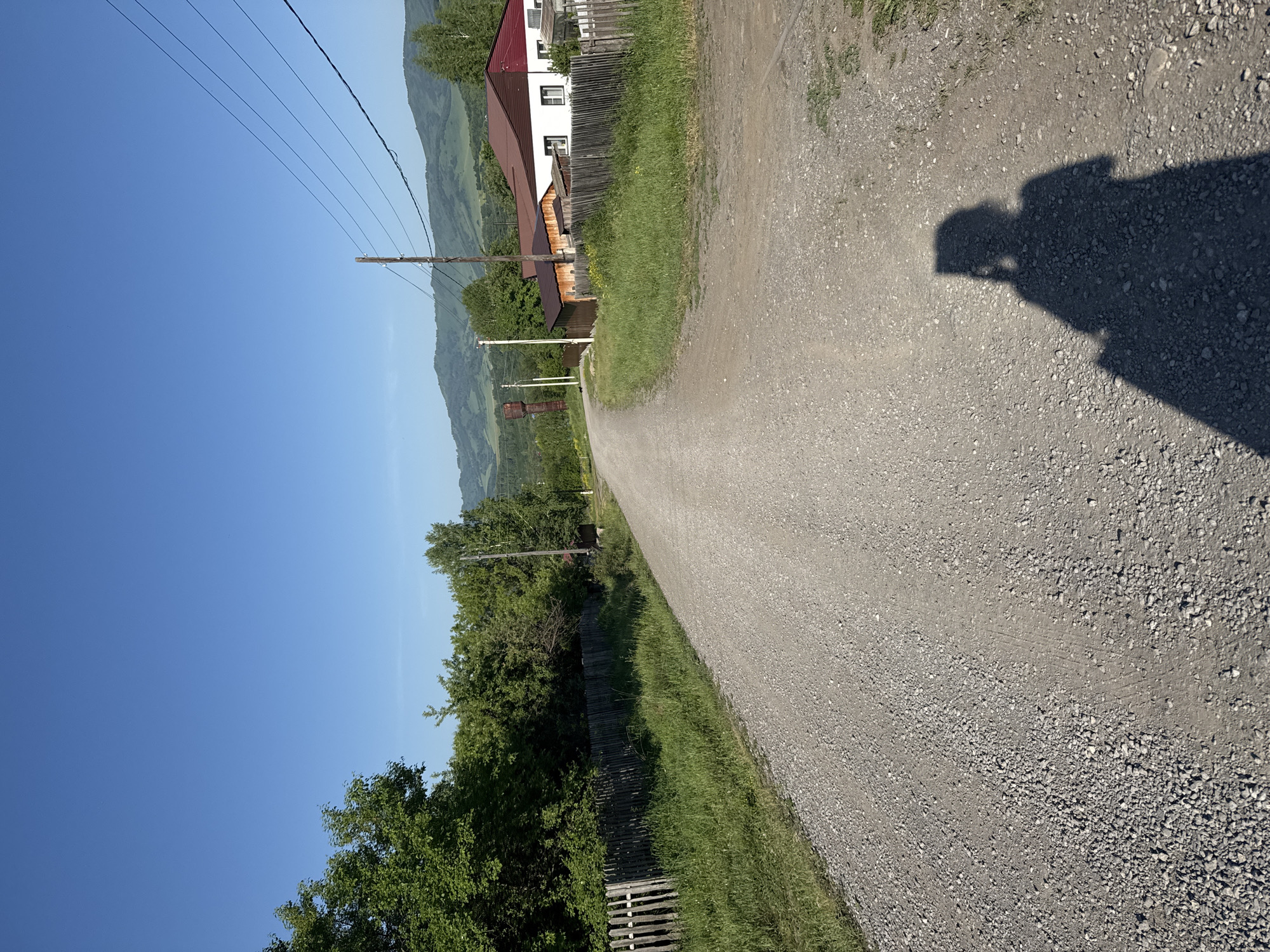 The image depicts a gravel road winding through a rural landscape under a clear blue sky. On the left side of the road, a weathered wooden fence runs alongside a grassy embankment and dense green trees. On the right side, houses with red roofs are visible, bordered by wooden fences and lush green grass. Power lines stretch across the scene, supported by wooden poles. In the distance, rolling green hills form the horizon. A prominent shadow of a person holding a phone is cast on the gravel road in the foreground, suggesting the photograph was taken by someone walking or standing on the road. The overall scene conveys a sense of a quiet, sun-drenched day in a village setting. The presence of power lines and houses indicates a populated area, likely a village or small town. The specific location of Soloneshnoye, Russia, is mentioned, placing the scene within a mountainous or hilly region.