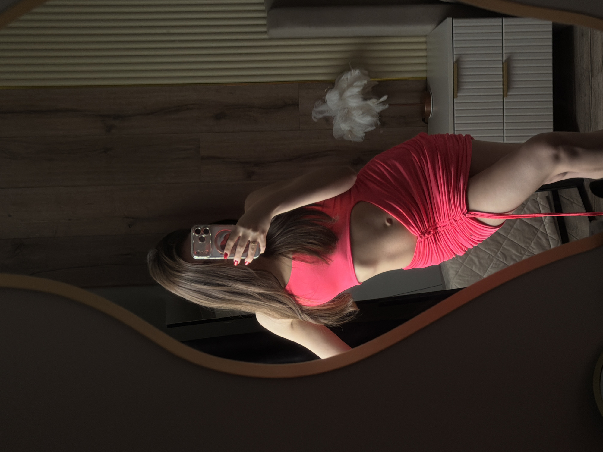 The image displays a mirror selfie taken by a woman indoors, with the provided location context of Chaikovsky, Russia. The woman, with long, light brown hair, is seen from her mid-torso to her upper legs, dressed in a vibrant pink or coral-colored, ruched mini dress or top that highlights her midriff. She is holding a silver smartphone, likely an iPhone with multiple cameras, in her right hand, obscuring her face in the reflection. Her fingernails are painted red.
The reflection captures a room interior, likely a bedroom or dressing area. In the background, there is a light-colored wall with horizontal slatted paneling. Below this, a wooden floor is visible, featuring a white, fluffy object that resembles a decorative item or a pet toy. To the right, a white bedside cabinet with two doors and horizontal ridged details is present, adorned with two gold or bronze-colored handles. A portion of a bed with a quilted, textured grey cover can also be seen. The mirror itself has a dark, possibly wooden, oval frame which partially frames the bottom and top of the image. The lighting appears to be artificial, typical of an indoor setting. No specific text is visible within the image.