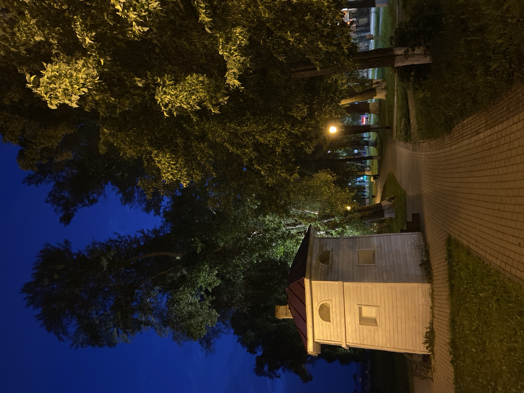 A small, quaint wooden building with a dark red roof stands beside a brick-paved pathway. The building has multiple square windows and arched window openings, suggesting it might be a small public structure like a ticket booth or a park office. It is surrounded by lush green grass and large trees, some of which are illuminated by a nearby streetlamp. The scene is set at night, with a deep blue sky visible above the trees, indicating it's likely dusk or early evening. The pathway curves away into the distance, lined with trees and more grassy areas. In the background, glimpses of buildings and streetlights suggest an urban or suburban setting. The overall atmosphere is peaceful and quiet, illuminated by artificial lights against the dark night. There is no visible text in the image.