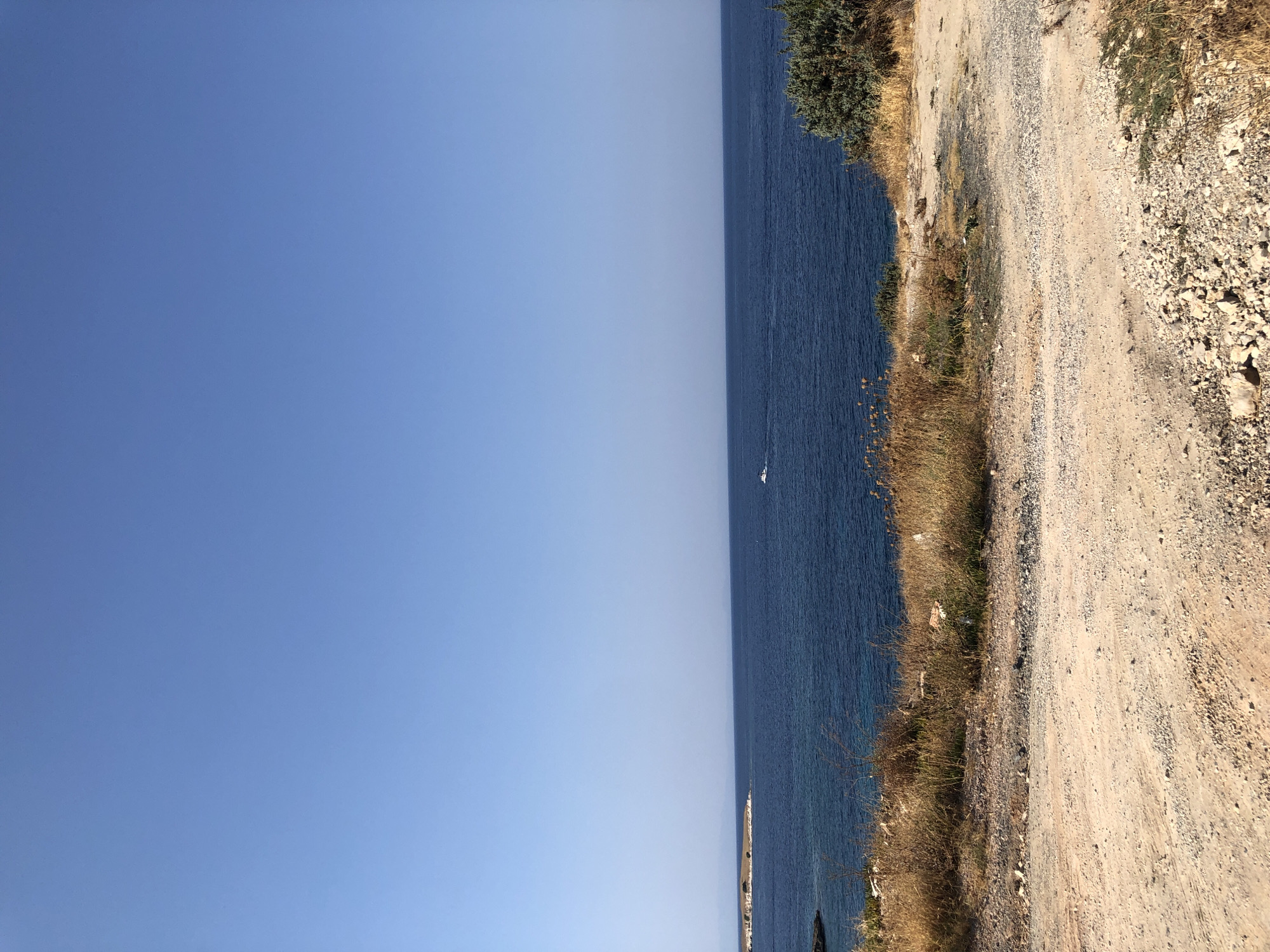 An unobstructed, clear blue sky dominates the upper portion of this landscape photograph. The horizon line is low, emphasizing the vastness of the sky. Below the sky, a deep blue ocean stretches horizontally across the frame. On the left side of the ocean, a small, rocky island with sparse vegetation is visible. In the middle distance, a white speedboat is moving across the water. The foreground features a dry, dusty, and rocky terrain, possibly a dirt road or path. Patches of dry, golden grass and scrubby bushes are scattered across this foreground area, suggesting a dry climate. The overall impression is of a coastal scene under a bright, sunny sky. The location is identified as Karaburun, Turkey.